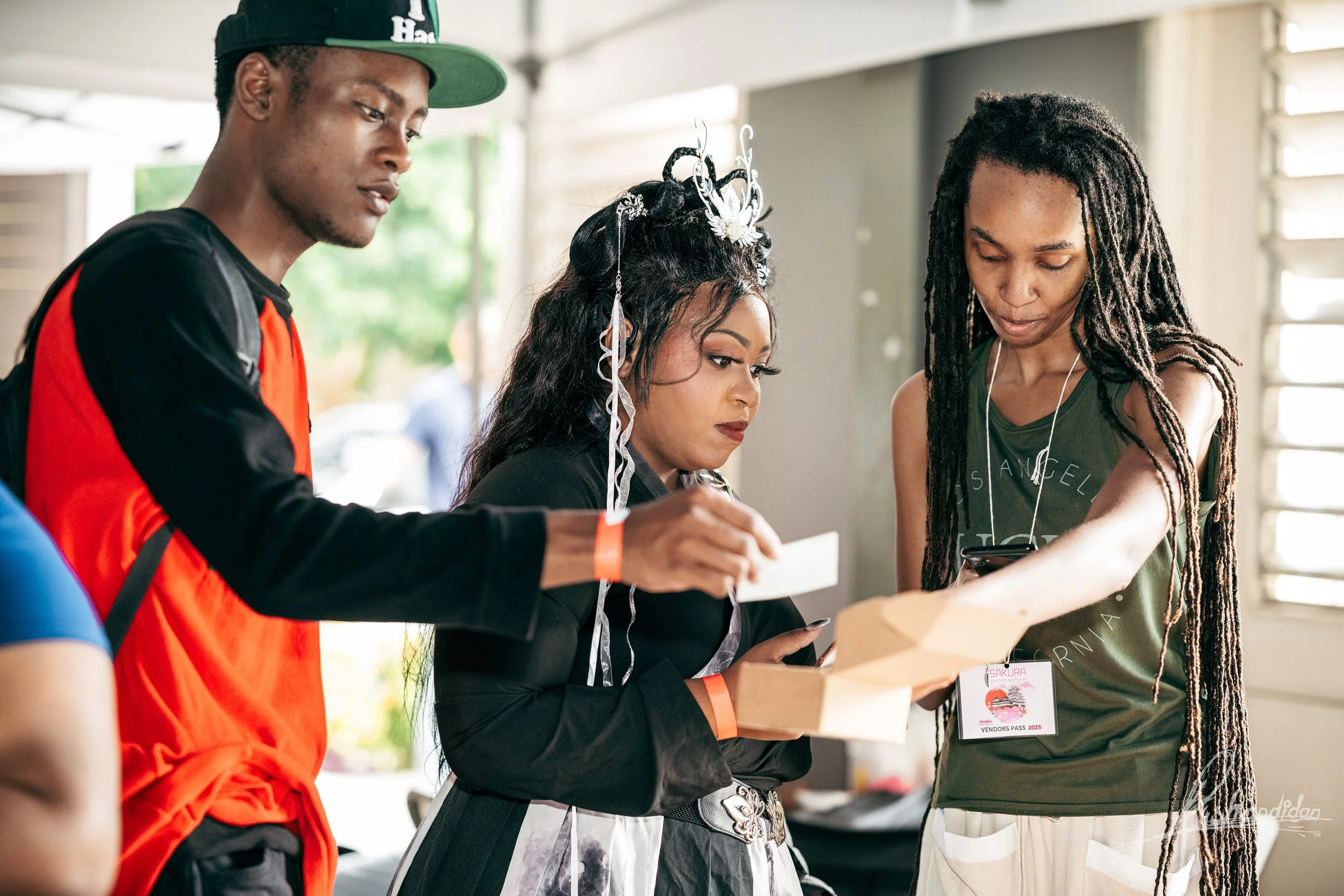 Three people gathered around looking at a box, one holding a paper, with a woman in the center wearing a crown and elaborate hairstyle.