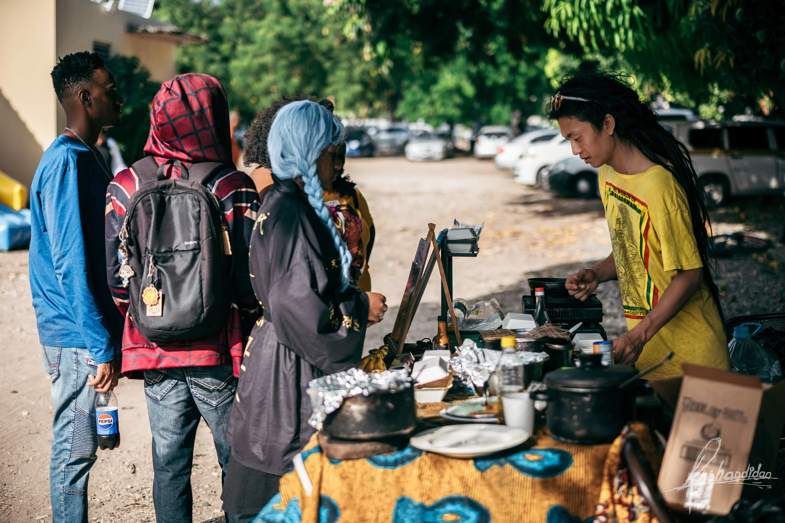 A diverse group of people, including a woman with long dark hair wearing a yellow shirt, stands at a food stall outdoors with parked cars and green trees in the background.