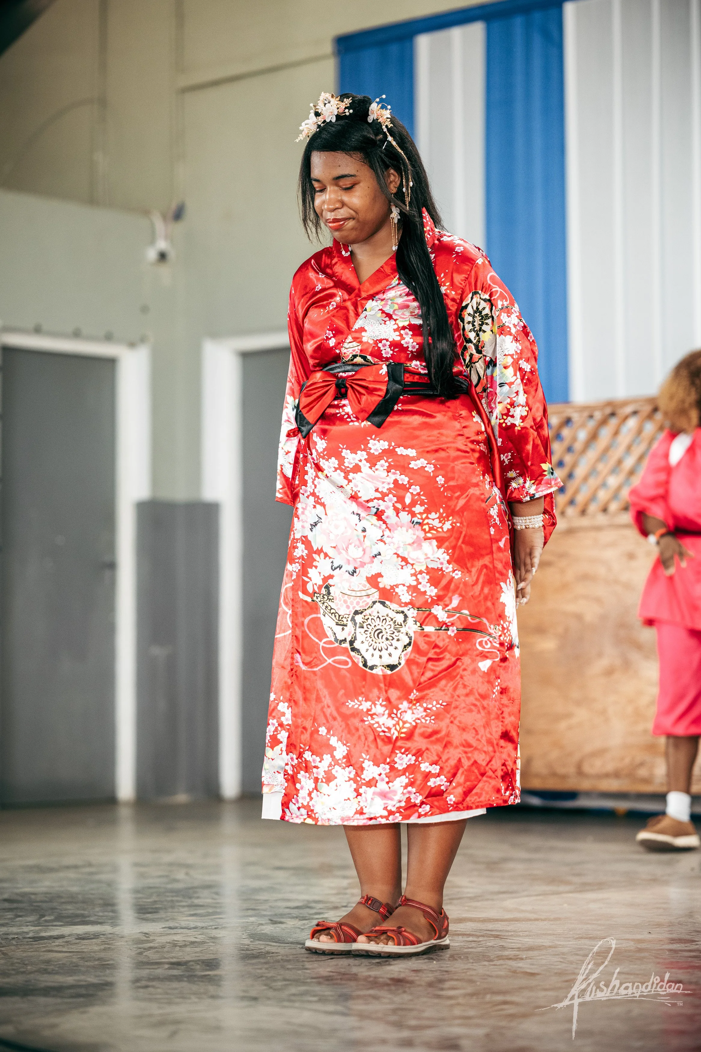 A woman dressed in a red floral kimono with a black bow, standing on a wooden floor with her eyes closed. She is wearing sandals and a flower crown, with a young girl in a pink outfit in the background.