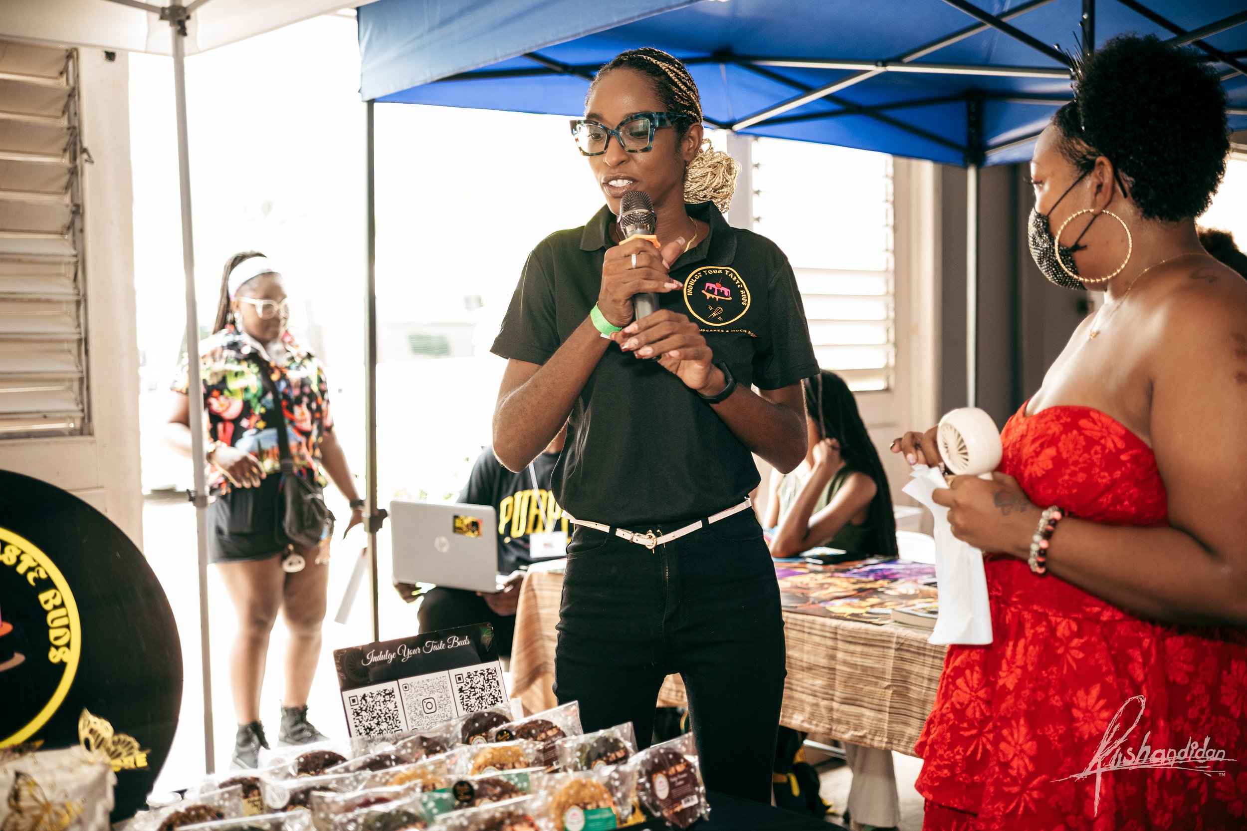A woman wearing glasses and a black polo shirt with a logo holds a microphone and speaks at an indoor event under a blue canopy. She is standing behind a table with packaged baked goods and a QR code card. Another woman in a red strapless dress and m