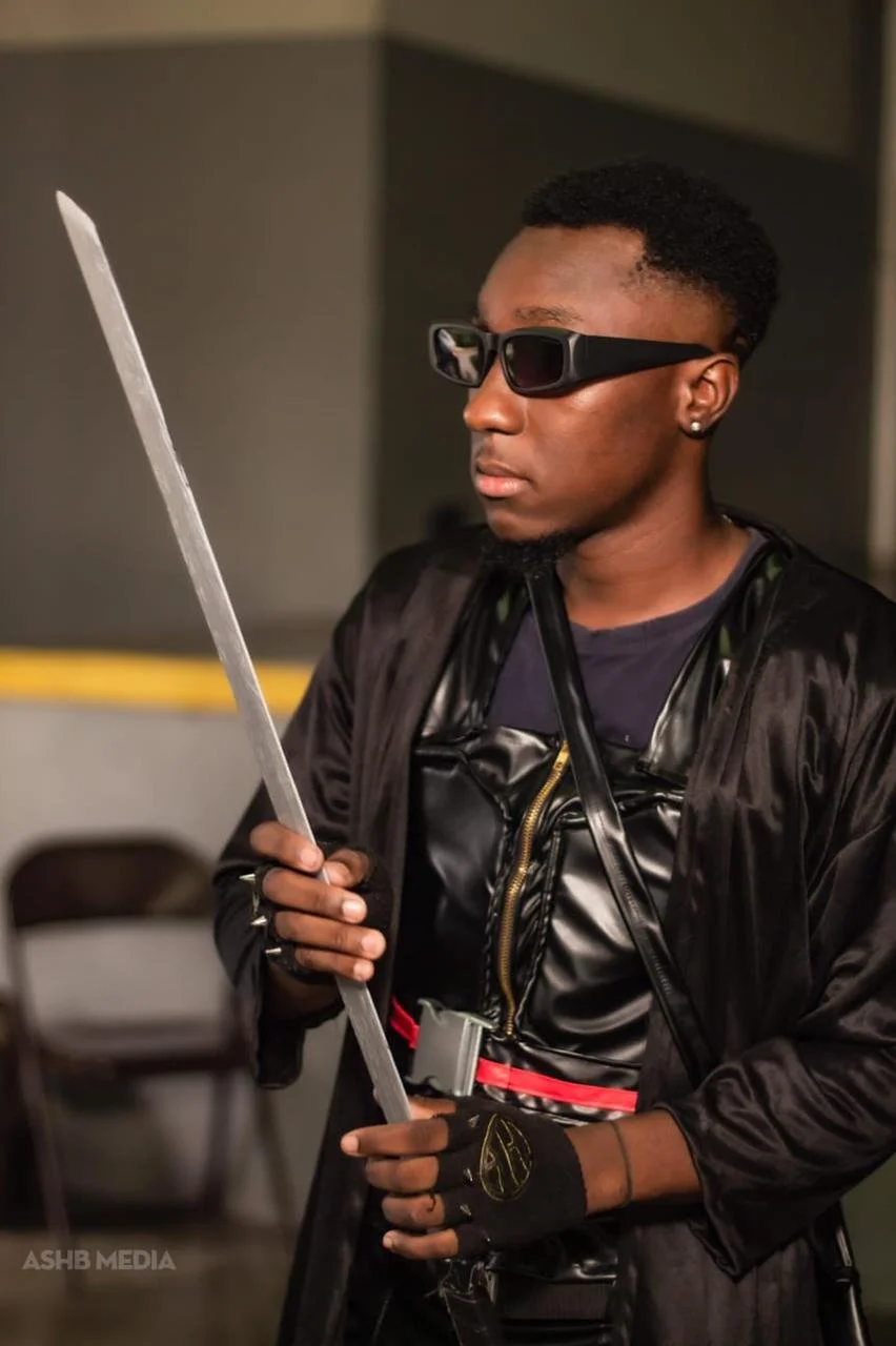 A young man wearing black sunglasses, black gloves, and a shiny black jacket, holding a long sword.