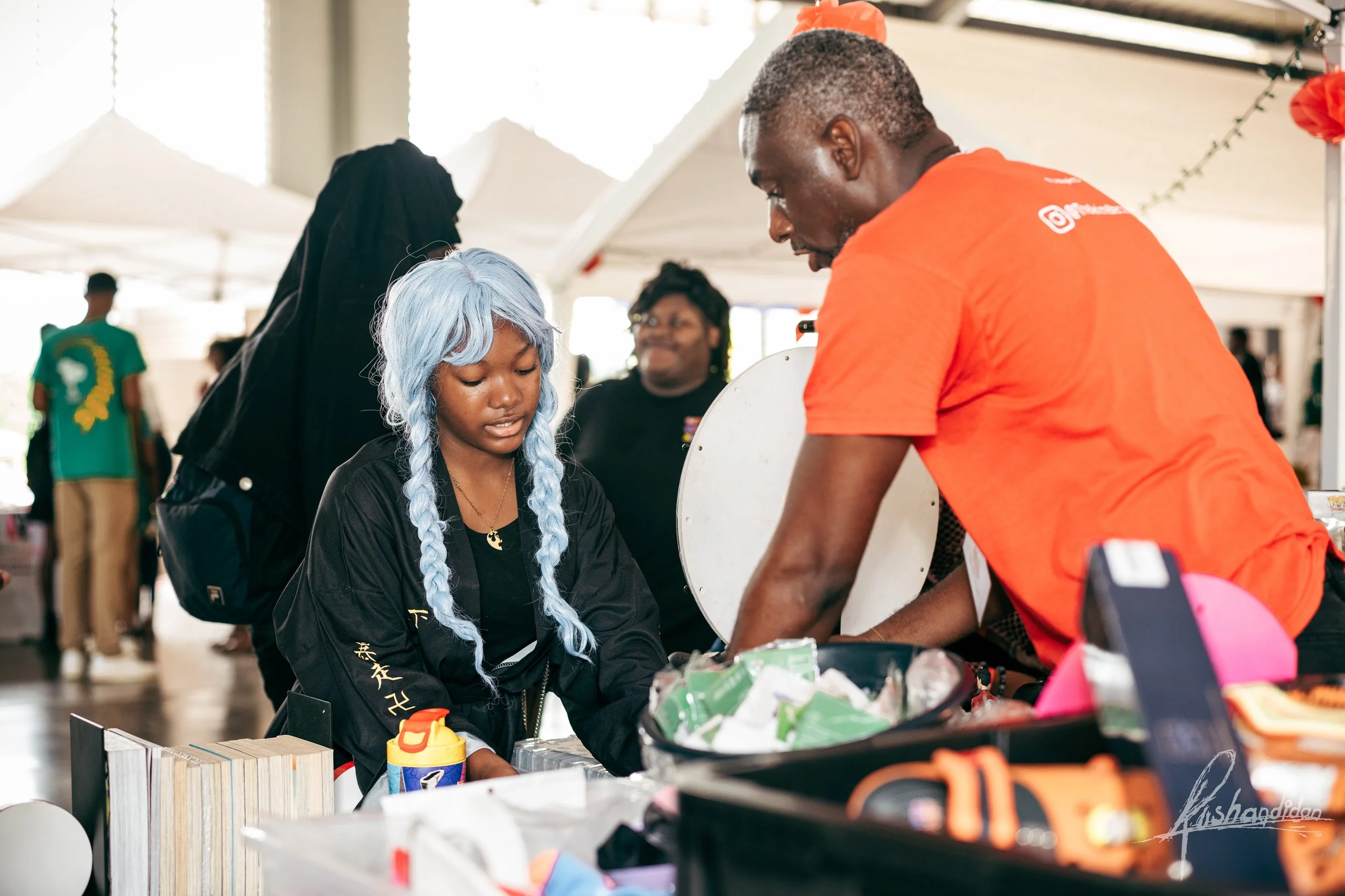 A young woman with light blue braided hair and black clothing shopping at a market stall, speaking with a man in an orange shirt who is behind the stall. In the background, there are other shoppers and white tents.