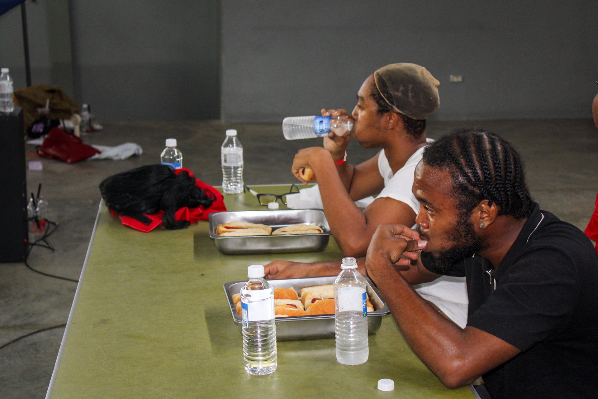 Two men sitting at a table with bottled water and trays of sandwiches. One man is drinking water while the other is resting his chin on his hand.
