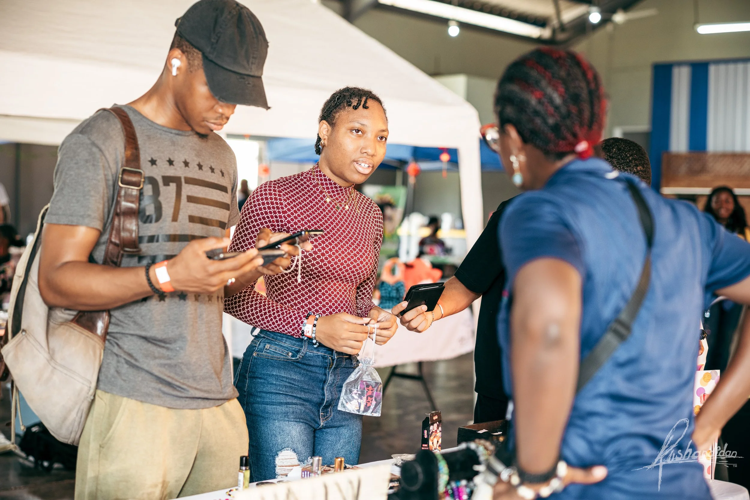 Three people at a market stall, one woman in a red patterned top explaining jewelry to two others, a man and a woman.