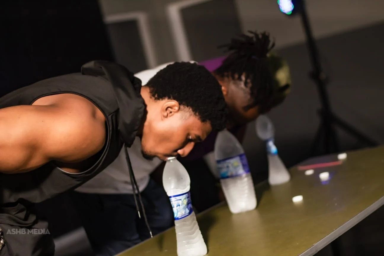 Two young men leaning over a table with water bottles, focused on something in front of them. The shirtless man wears black athletic apparel, and the other has dreadlocks and a gray shirt.