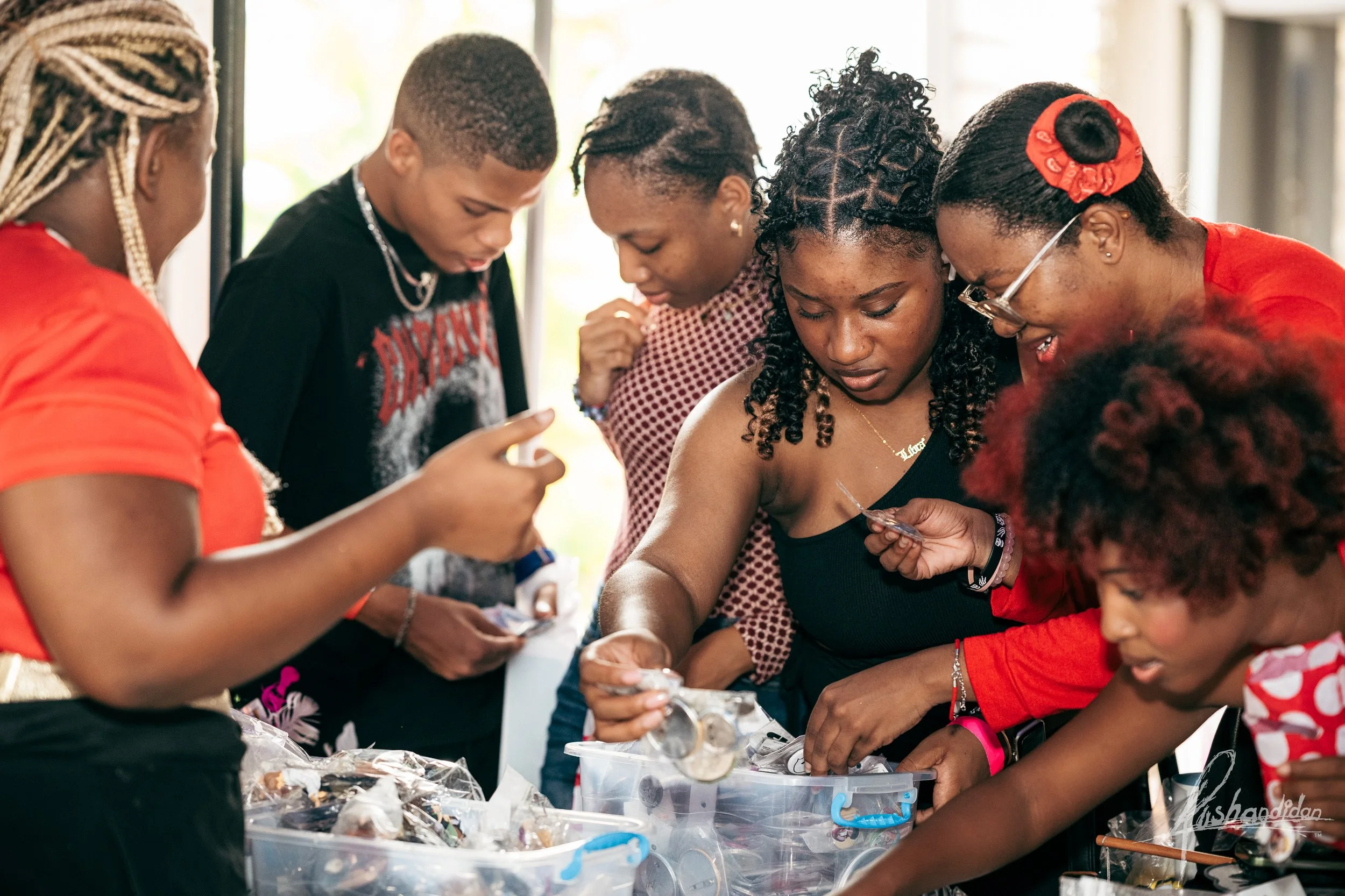 A group of six people, including children and adults, gathered around a table, inspecting and handling containers of coins and small objects.