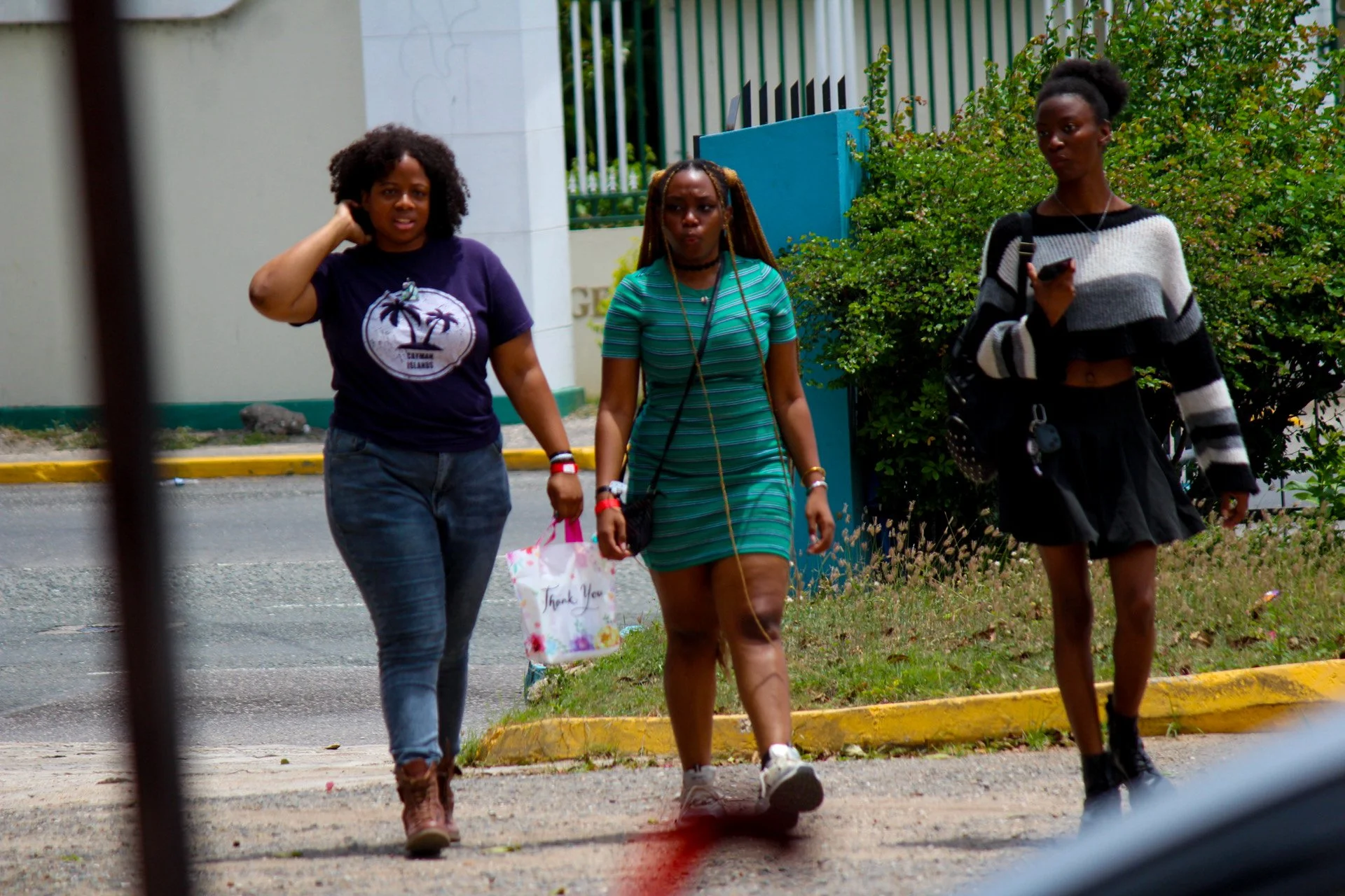 Three young women walking outdoors on a sunny day, with trees and a building in the background.
