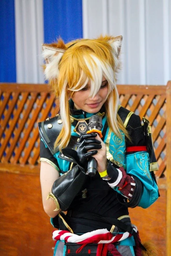 Person dressed in a cosplay costume with fox ears, holding a microphone, standing in front of a wooden lattice background.