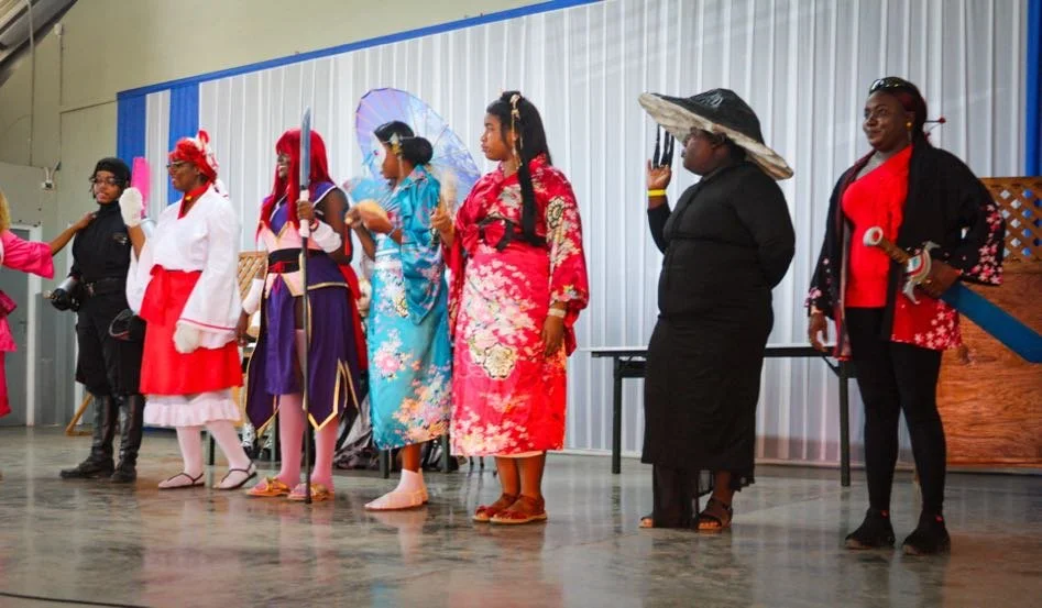 A group of people dressed in various costumes, standing on a stage, possibly during a cosplay or themed event.