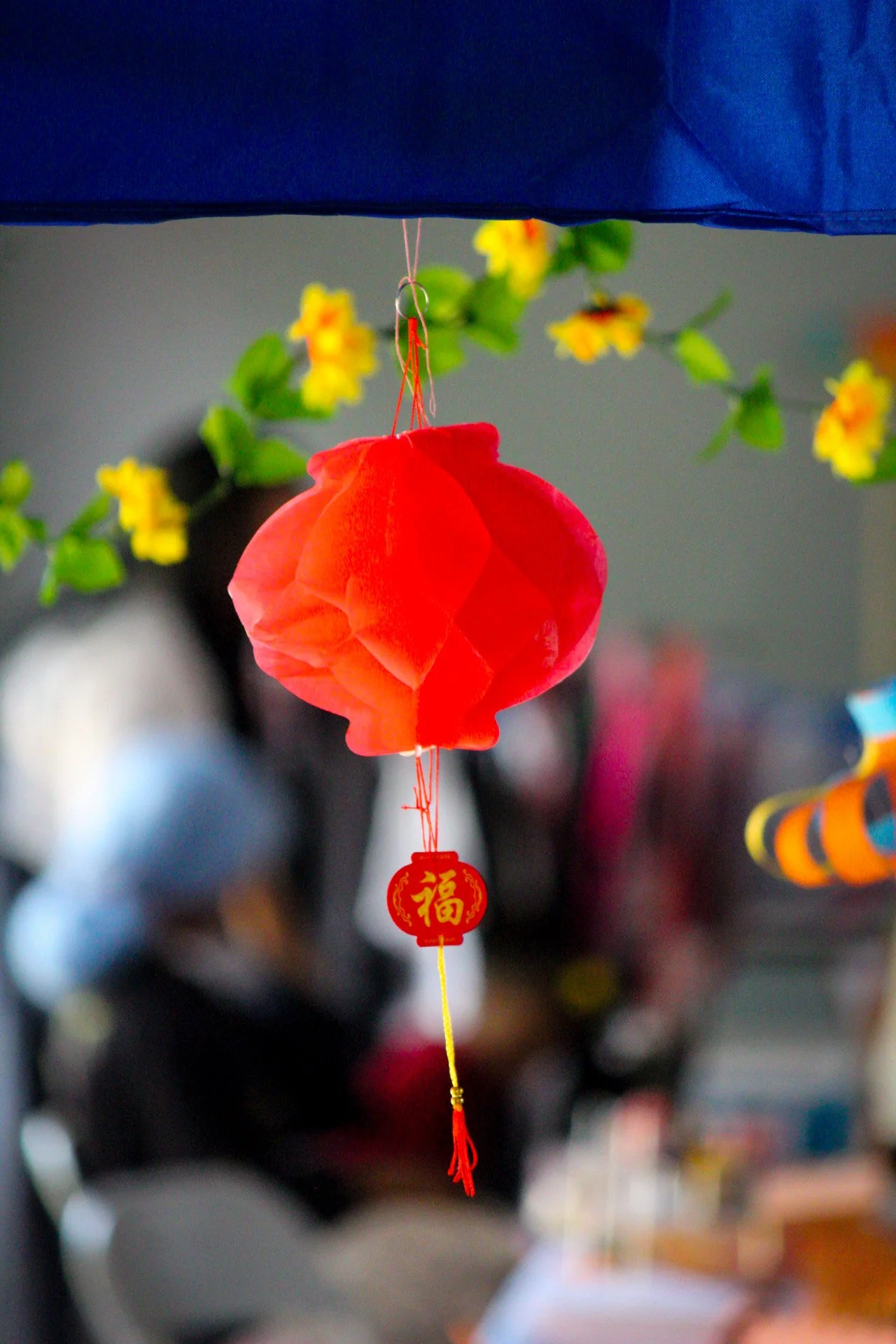 Red paper lantern with a yellow Chinese character, hanging from a string decorated with a small red and gold emblem, amidst yellow flowers and blurred background.