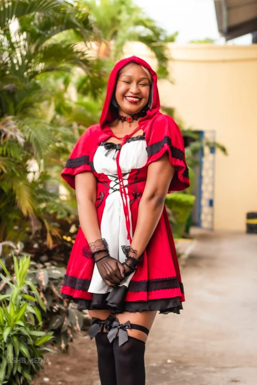 Woman dressed as Little Red Riding Hood standing outdoors, smiling with eyes closed. She wears a red hooded cape, black lace gloves, a white and black dress with red accents, and thigh-high black stockings with bows.