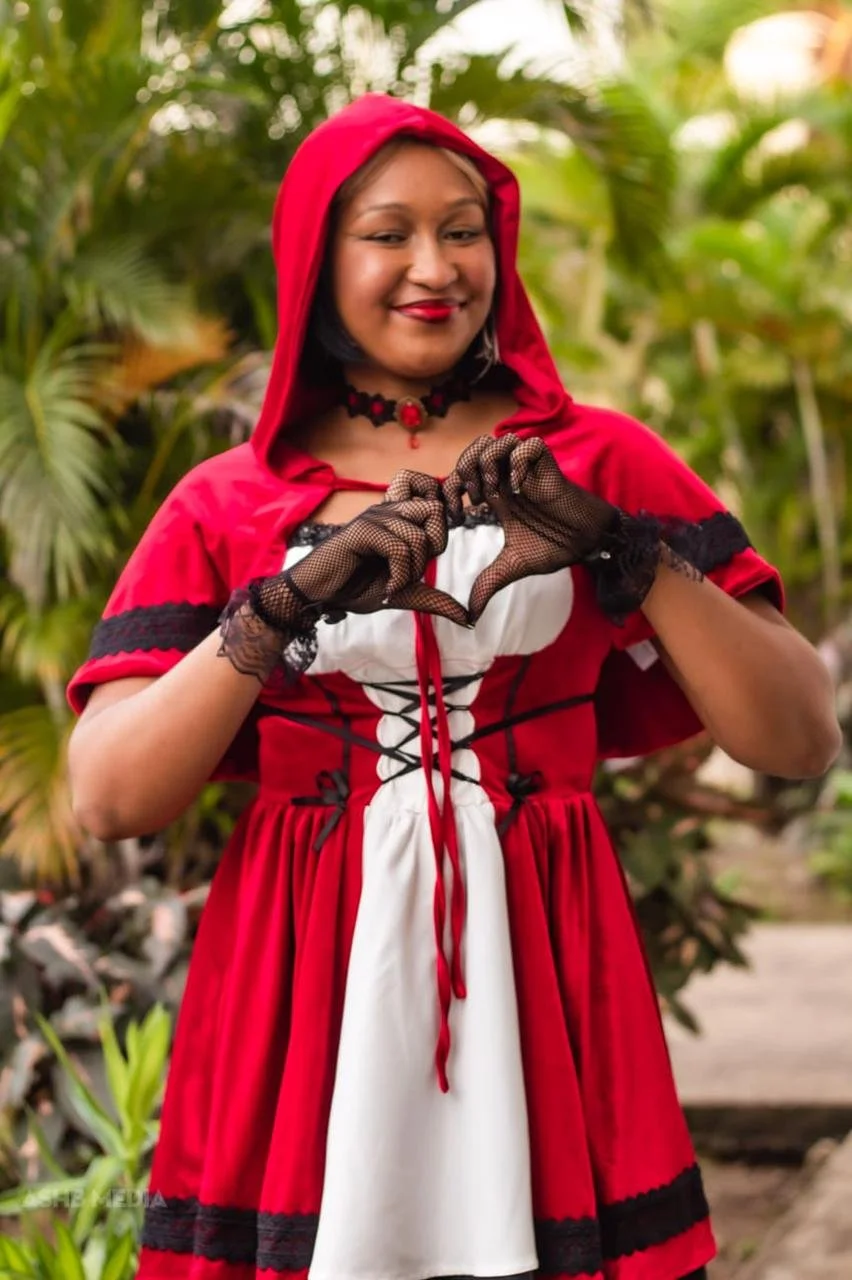 A woman dressed as Little Red Riding Hood making a heart shape with her hands, standing outdoors with green foliage in the background.