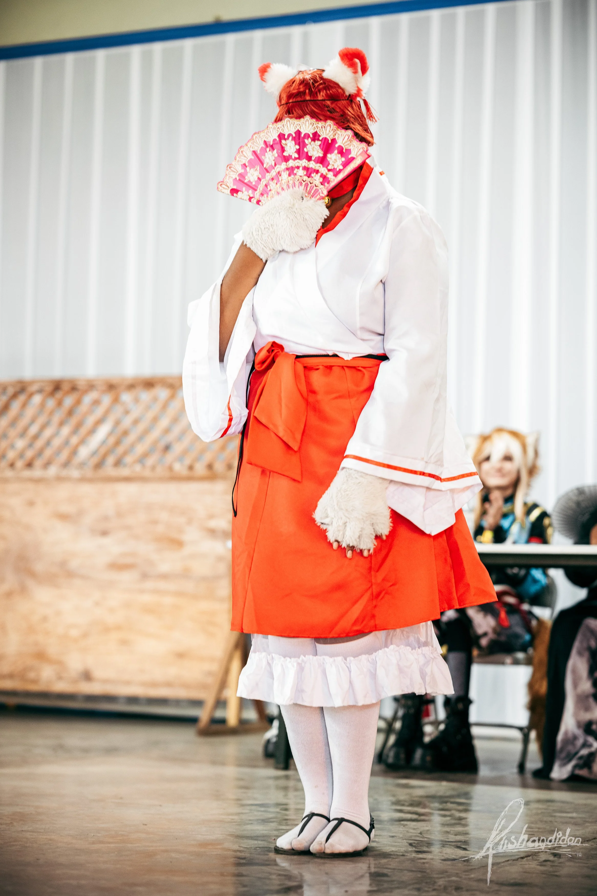 Person dressed in a cosplay costume of a cat character, wearing a kimono-style outfit, holding a decorative pink folding fan, with cat ears headband, and standing on a wooden floor in an indoor setting.