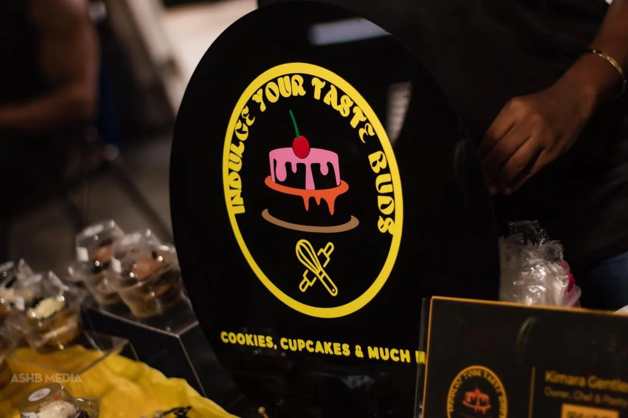 Black circular sign with yellow text that reads, "Indulge Your Taste Buds" and "Cookies, Cupcakes & Much More". The sign features a pink cupcake with blue icing and a red cherry on top, with an orange ring beneath it, and two crossed baking tools at 