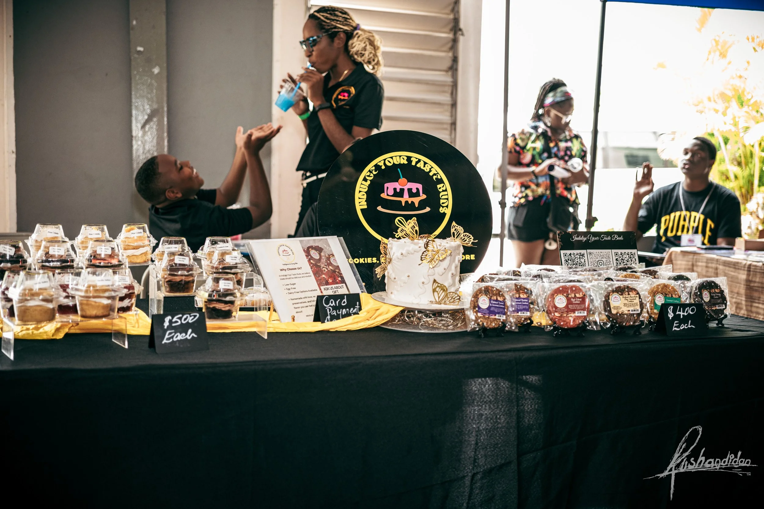A table at a bakery or dessert stand with various cupcakes and cookies displayed for sale. Several people are present, including a woman speaking to a young boy, two women in the background, and a man sitting at the table, gesturing. There is a cake 