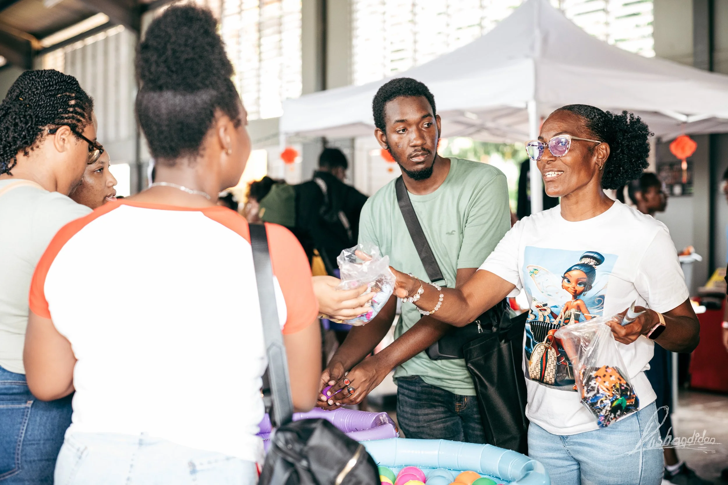 A woman selling small toys or candies at a market stall to a group of young women, with a white tent and market setting in the background.