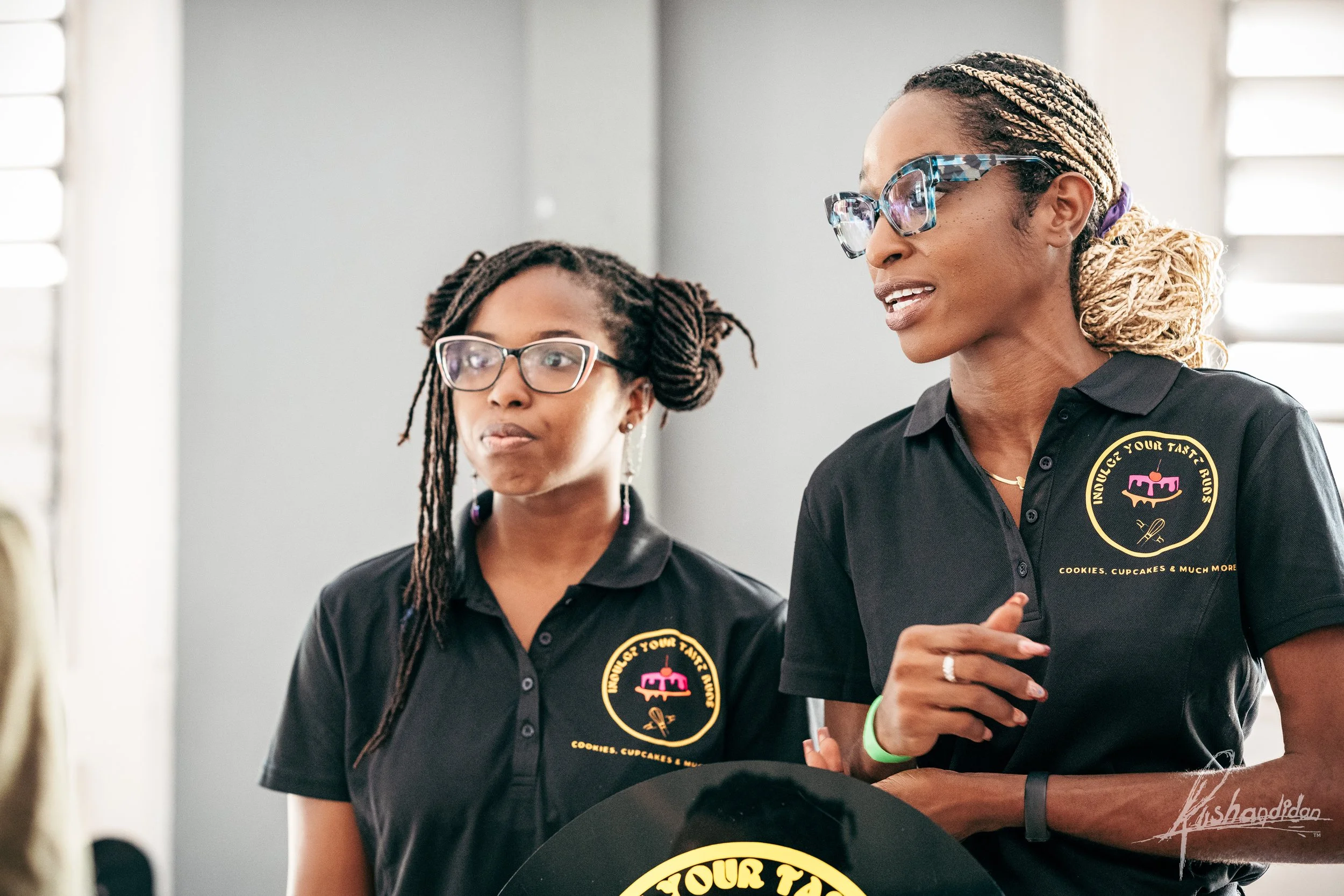 Two women wearing black polo shirts with a logo that reads "Indulge Your Taste Buds" are engaged in conversation indoors, with one woman holding a pen.