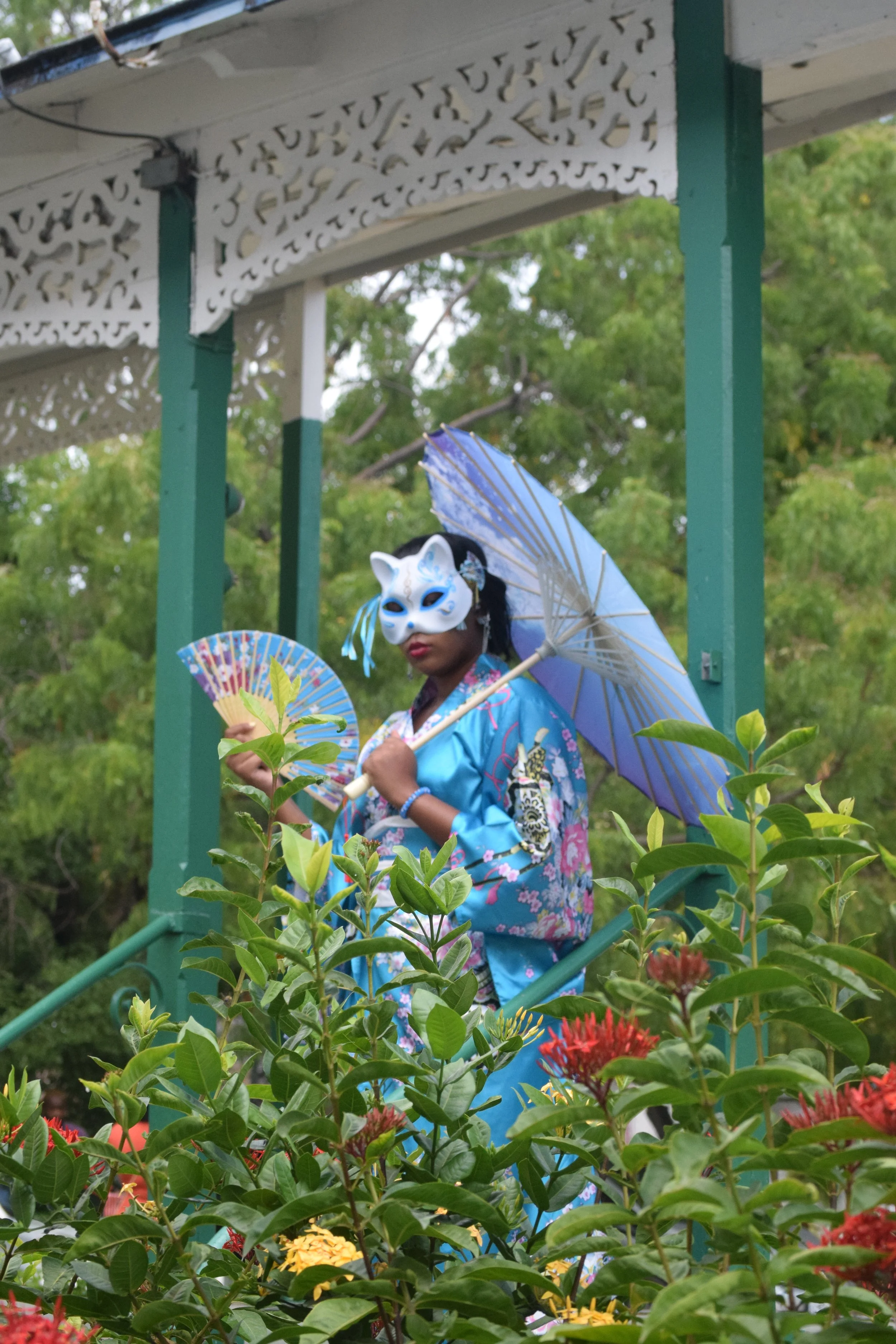 Person wearing a traditional Asian costume with a fox mask, holding a fan and an umbrella, standing on a green balcony with plants and flowers in the foreground.