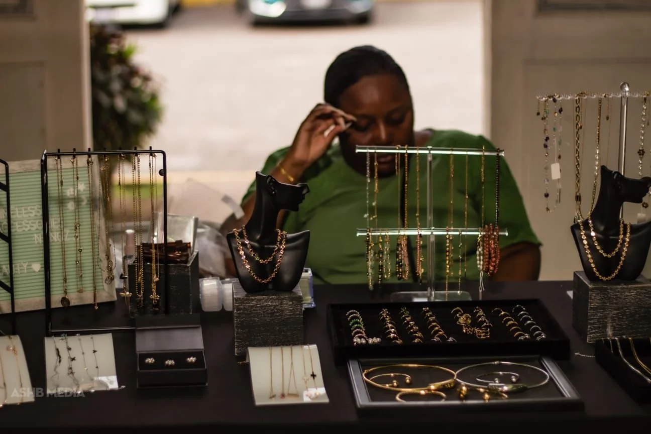 A woman shopping for jewelry at an outdoor market stall, with necklaces, earrings, and bracelets displayed on table and jewelry busts.