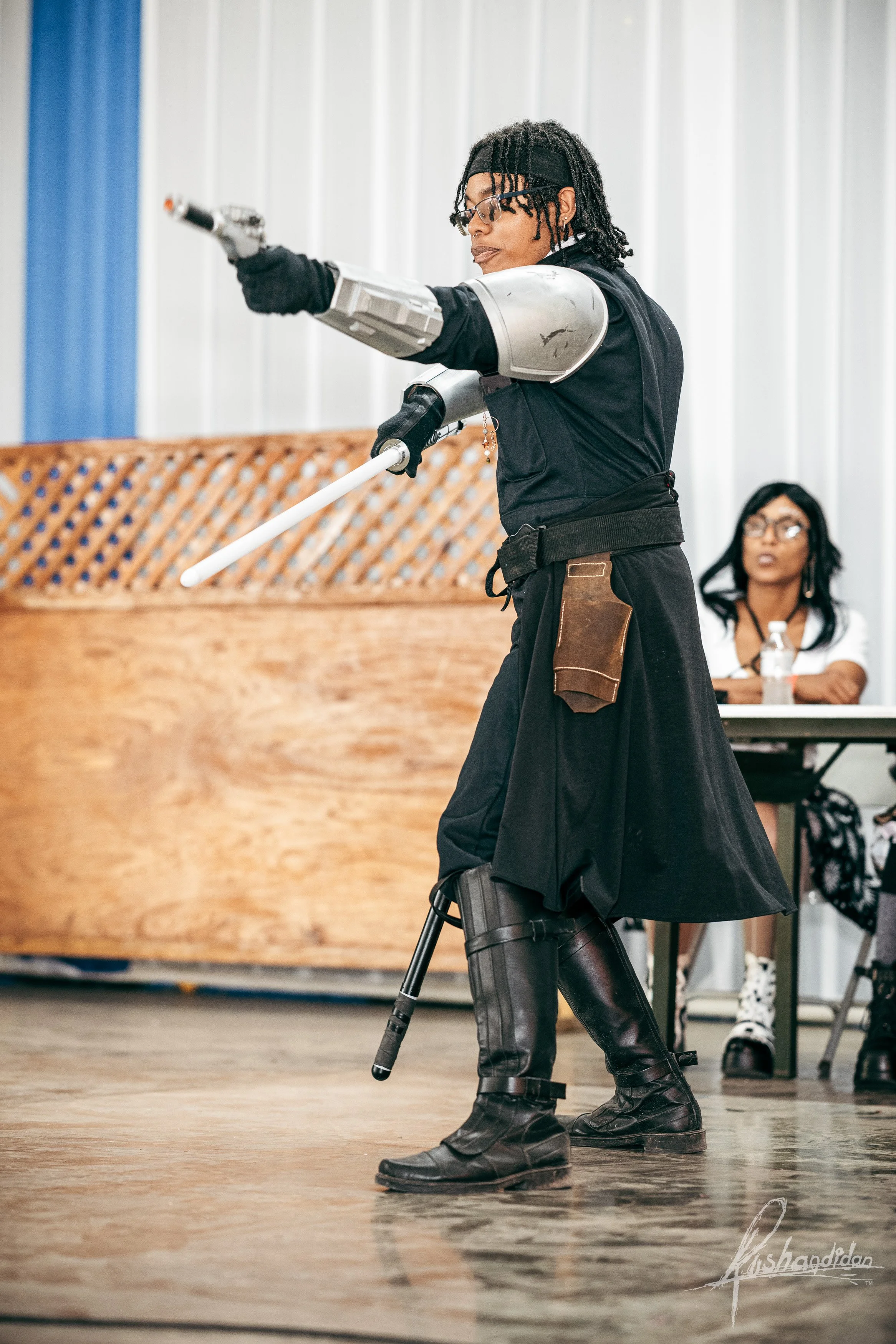 Person dressed as a futuristic warrior with robotic arm and leg, holding a sword, performing on a stage in front of an audience, with another person sitting at a table watching.