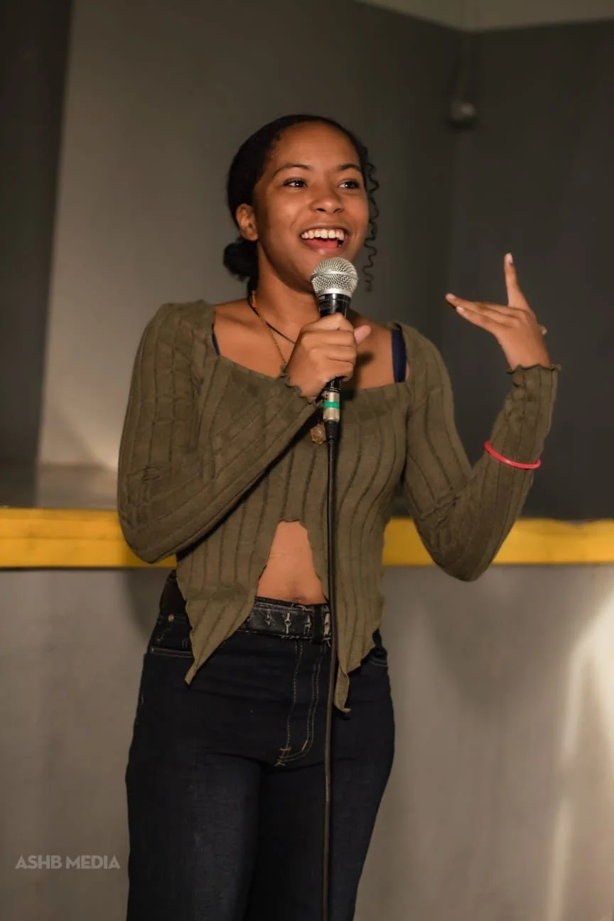 A young woman with dark hair tied back, smiling and speaking into a microphone, wearing a green cropped hoodie and black jeans, standing on a stage. 