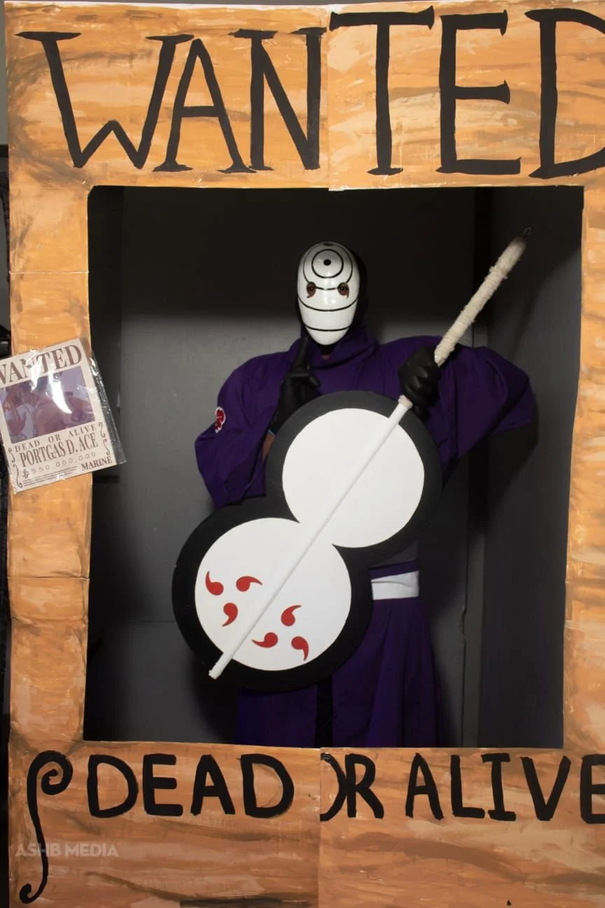 Costume display mimicking a Wanted poster, featuring a figure with a white mask with black spiral and dot design, dressed in purple, holding a black and white circular sign with red designs and a staff, surrounded by a wooden frame with the words "WA