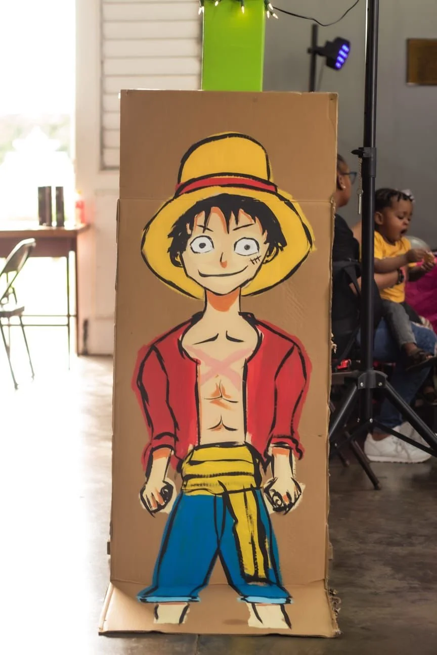 Hand-painted cartoon of a character with black messy hair, a yellow straw hat with a red band, a red open shirt, blue pants, and a yellow sash, holding two small objects, displayed on a cardboard stand.