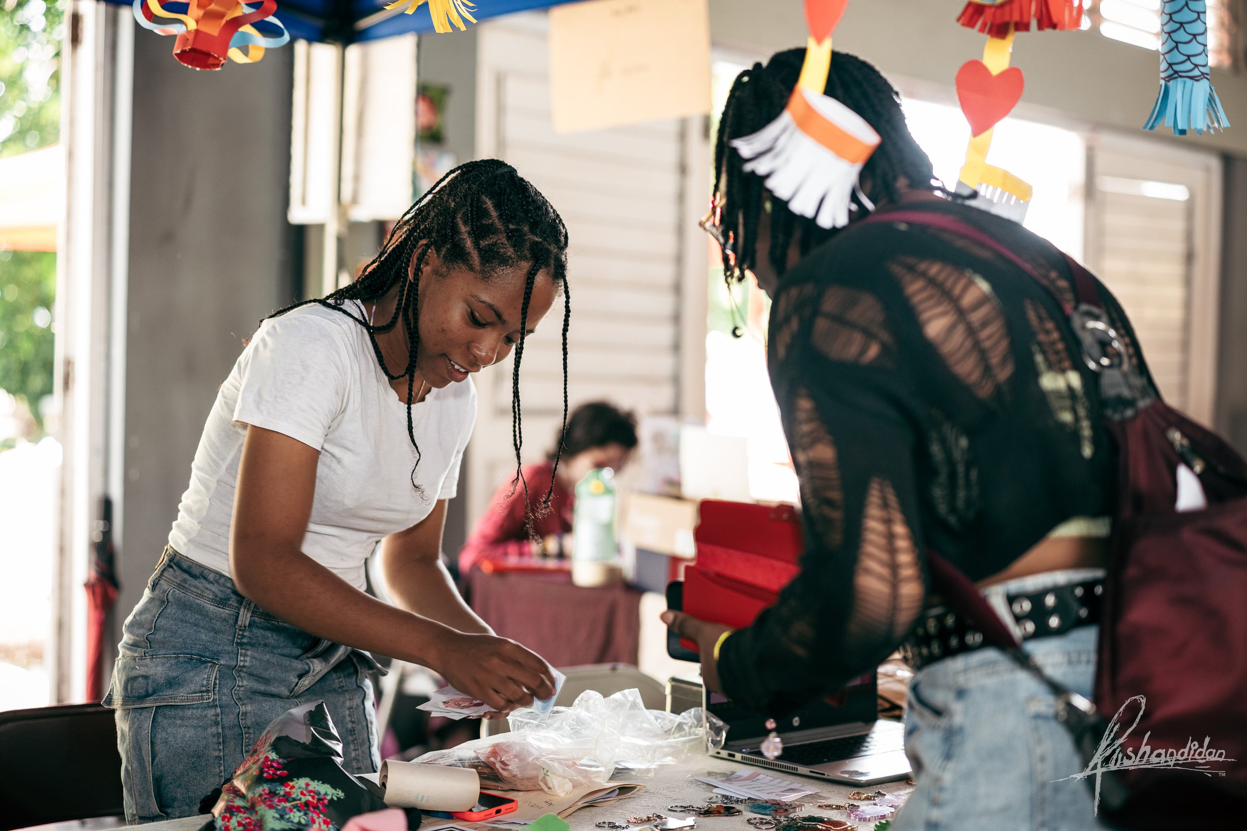 Two young women are at a table, looking at various craft items and papers, with colorful decorations hanging above them.