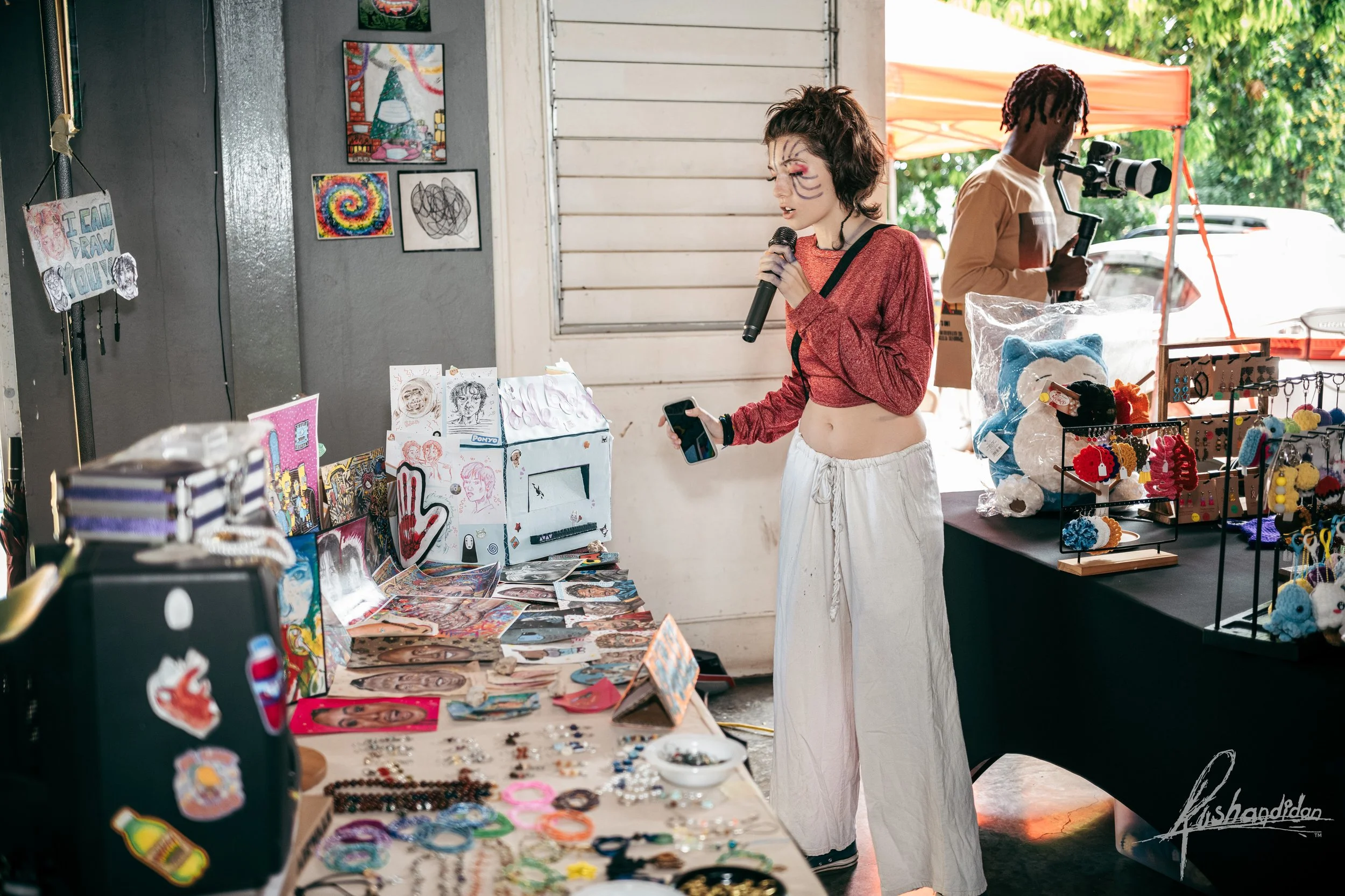 A young woman with painted face acting as a clown or performer, holding a microphone and a phone, standing at a vendor booth selling arts and crafts, with a person in the background taking photos with a camera, outside under a canopy.