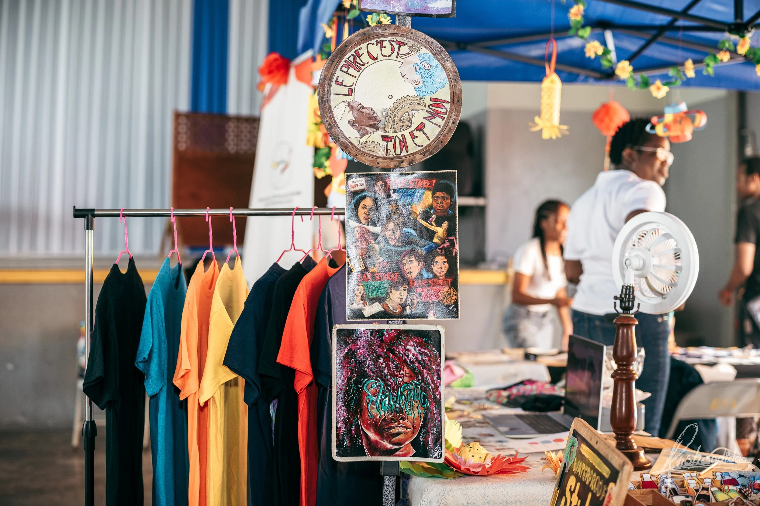 Indoor market stall with colorful T-shirts hanging on a rack, framed artwork and posters, and people browsing in the background.