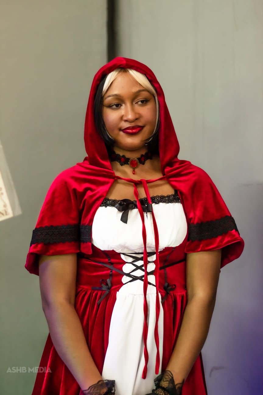 A woman dressed in a red hooded cape with black lace accents, a white and black dress with lace-up details, wearing black lace gloves and dark lipstick, standing against a plain wall.