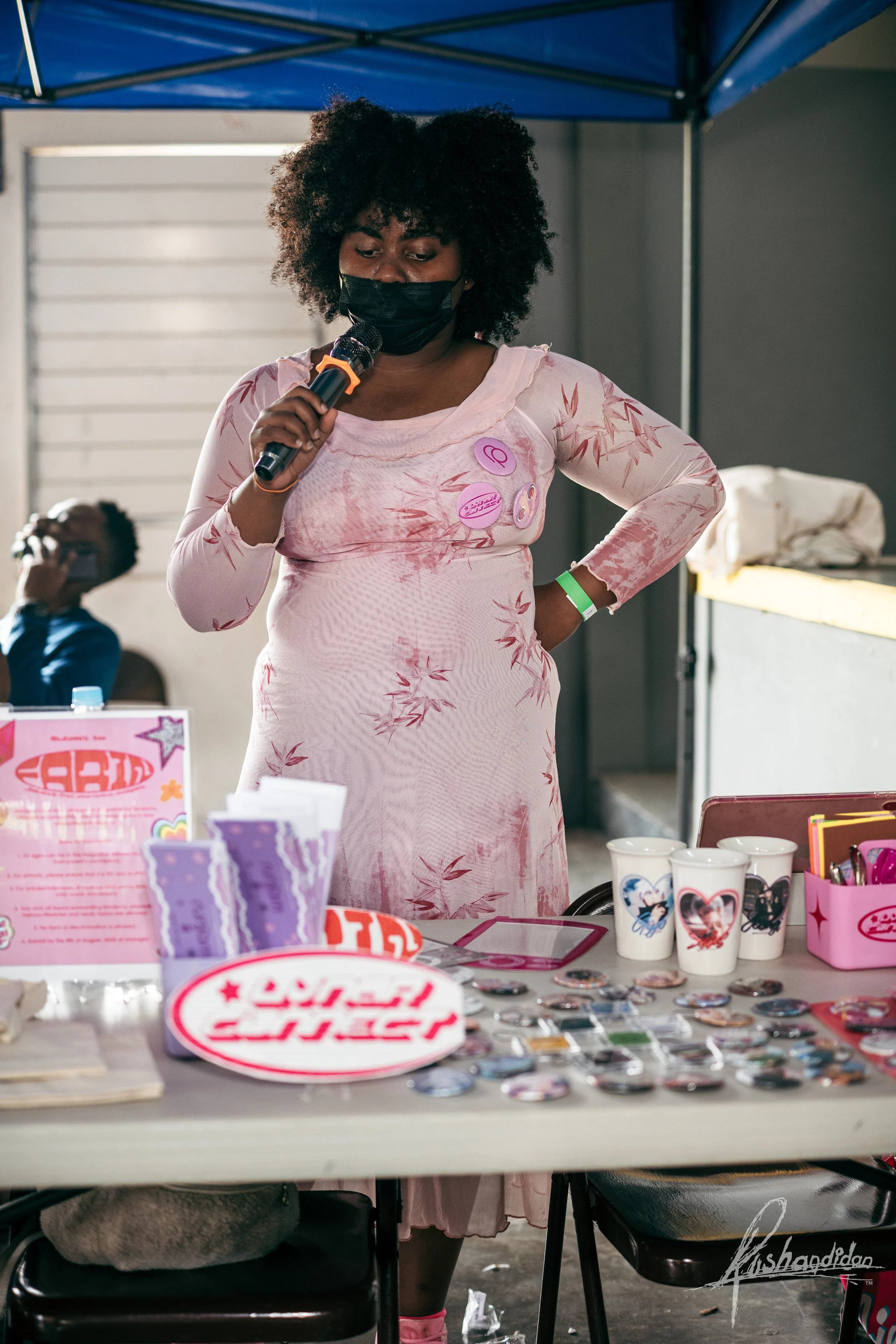 A woman with curly hair wearing a pink dress and a black face mask stands at a table, holding a microphone, at an event under a blue canopy. The table has various stickers, buttons, cups, and pink-themed items.