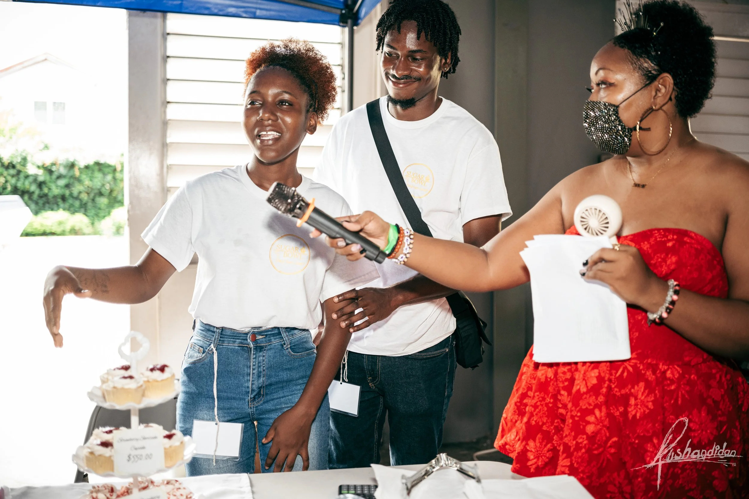 A woman in a red dress and face mask holding a microphone is interviewing a young girl who is speaking, with a man beside her. They are at a cake auction or sale, with cakes and price tags on the table.