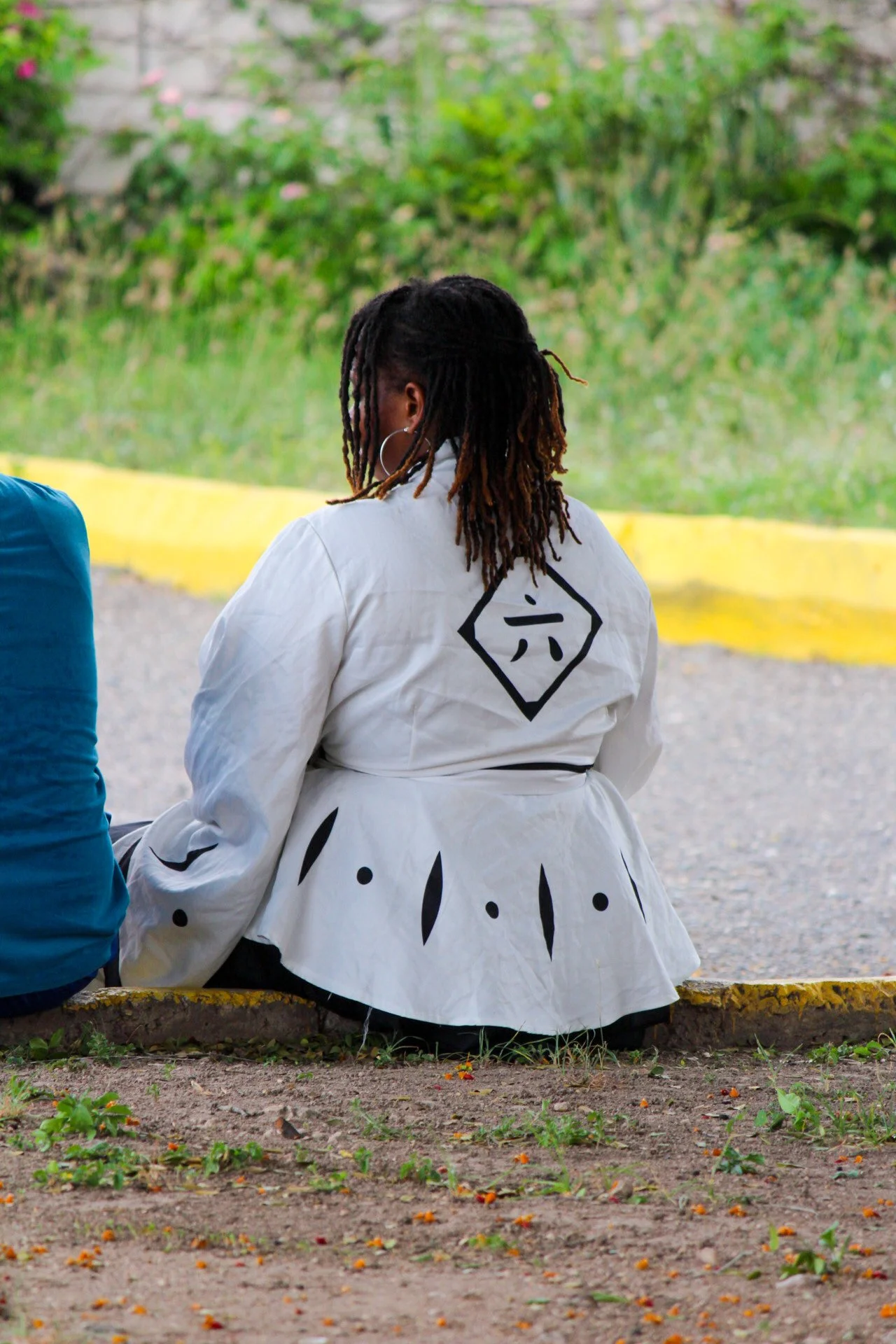 A woman with decorated dreadlocks sitting on the ground next to another person, outdoors with greenery and a yellow curb behind her.