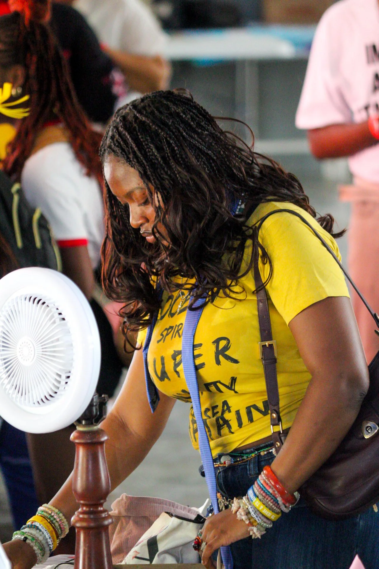 A woman in a yellow T-shirt with black writing, wearing multiple colorful bracelets, and with long curly hair, is bending over a table near a small white fan, in a busy indoor setting.