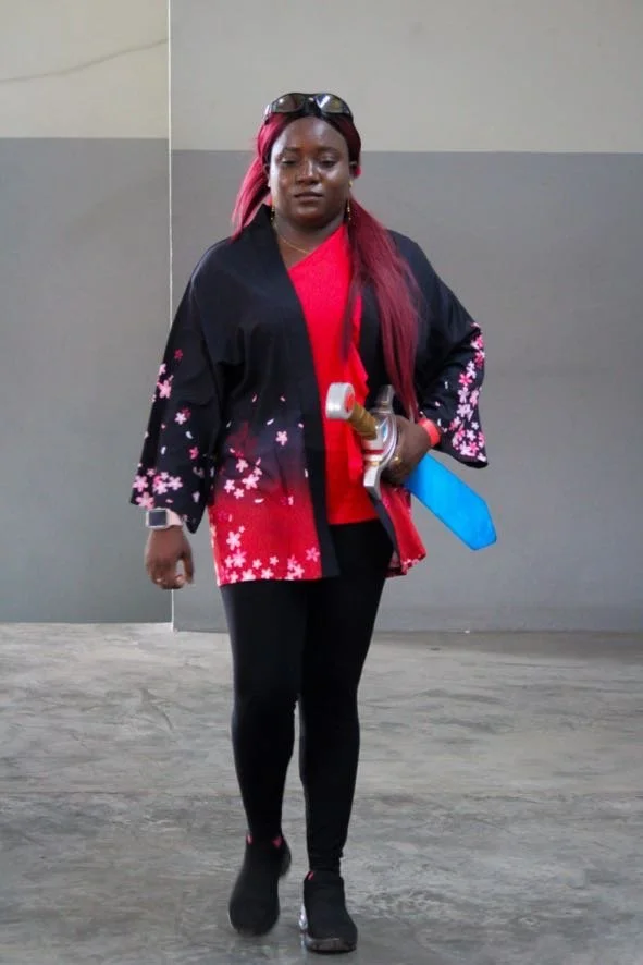 A woman with pink hair, wearing sunglasses on her head, a black jacket with pink and white floral patterns, a red shirt, black leggings, and boots, holding a sword and a hammer.