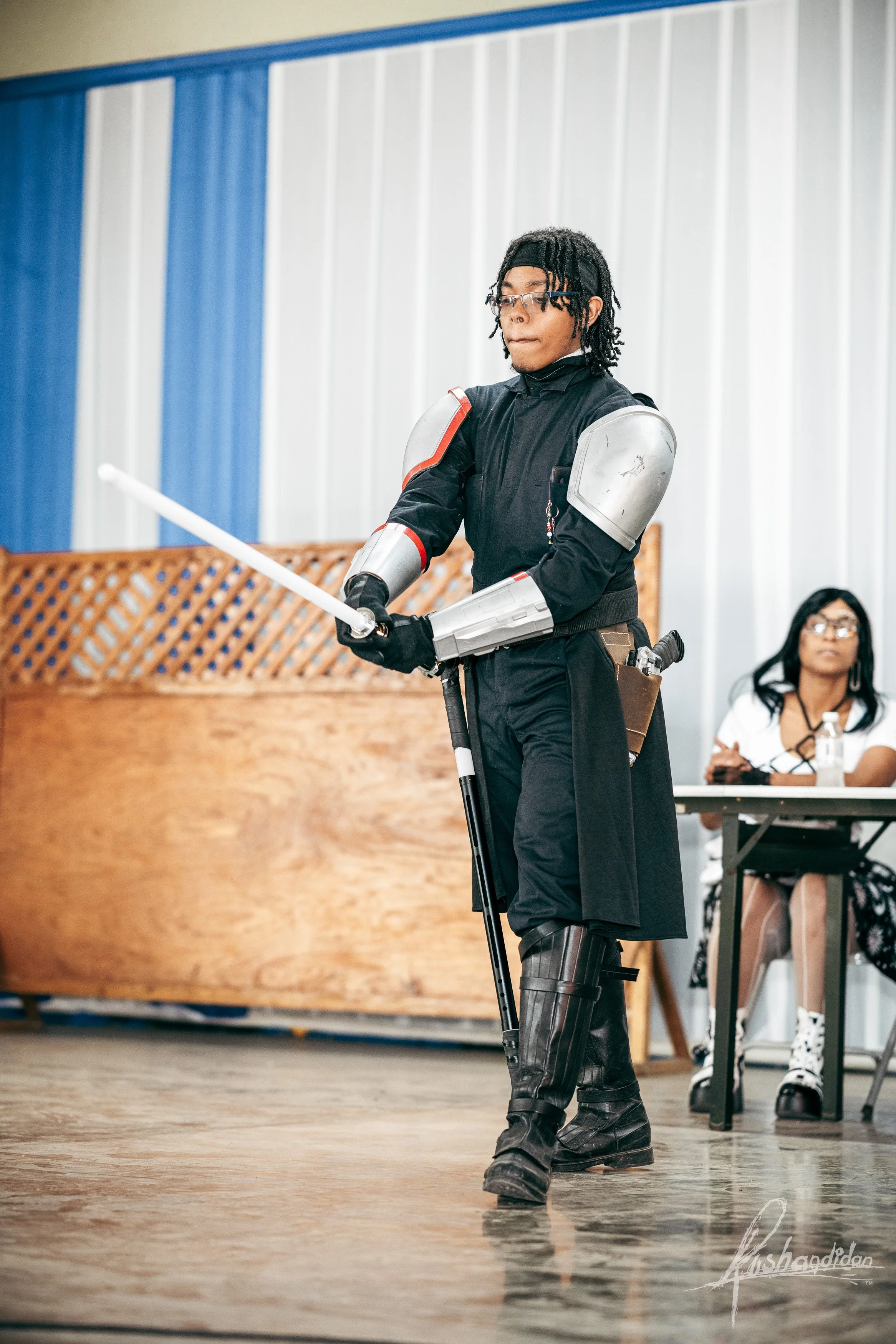 Person dressed as a futuristic knight in black armor with metallic shoulder and arm pieces, holding a white foam sword, standing in a room with blue and white walls, with a woman seated at a table in the background.