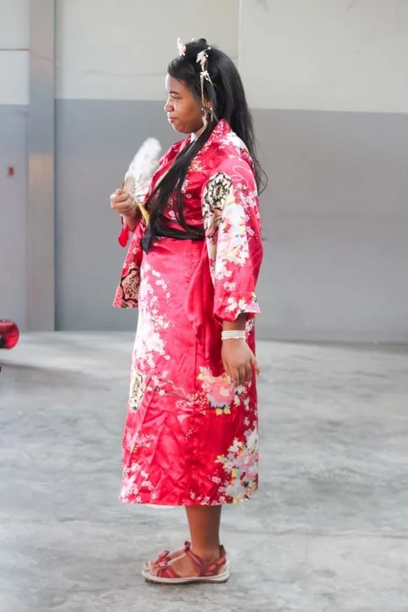 Young girl wearing a traditional red kimono with floral patterns, holding a paper fan, standing in a parking garage.