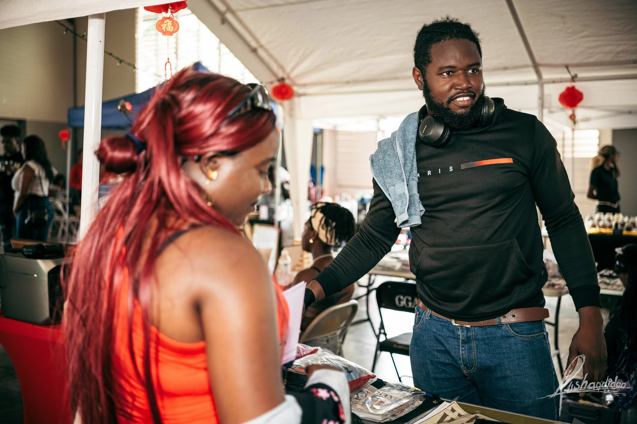 A man with headphones around his neck and a towel on his shoulder talks to a woman with red hair at an indoor market or event, with tables, chairs, and other people in the background.
