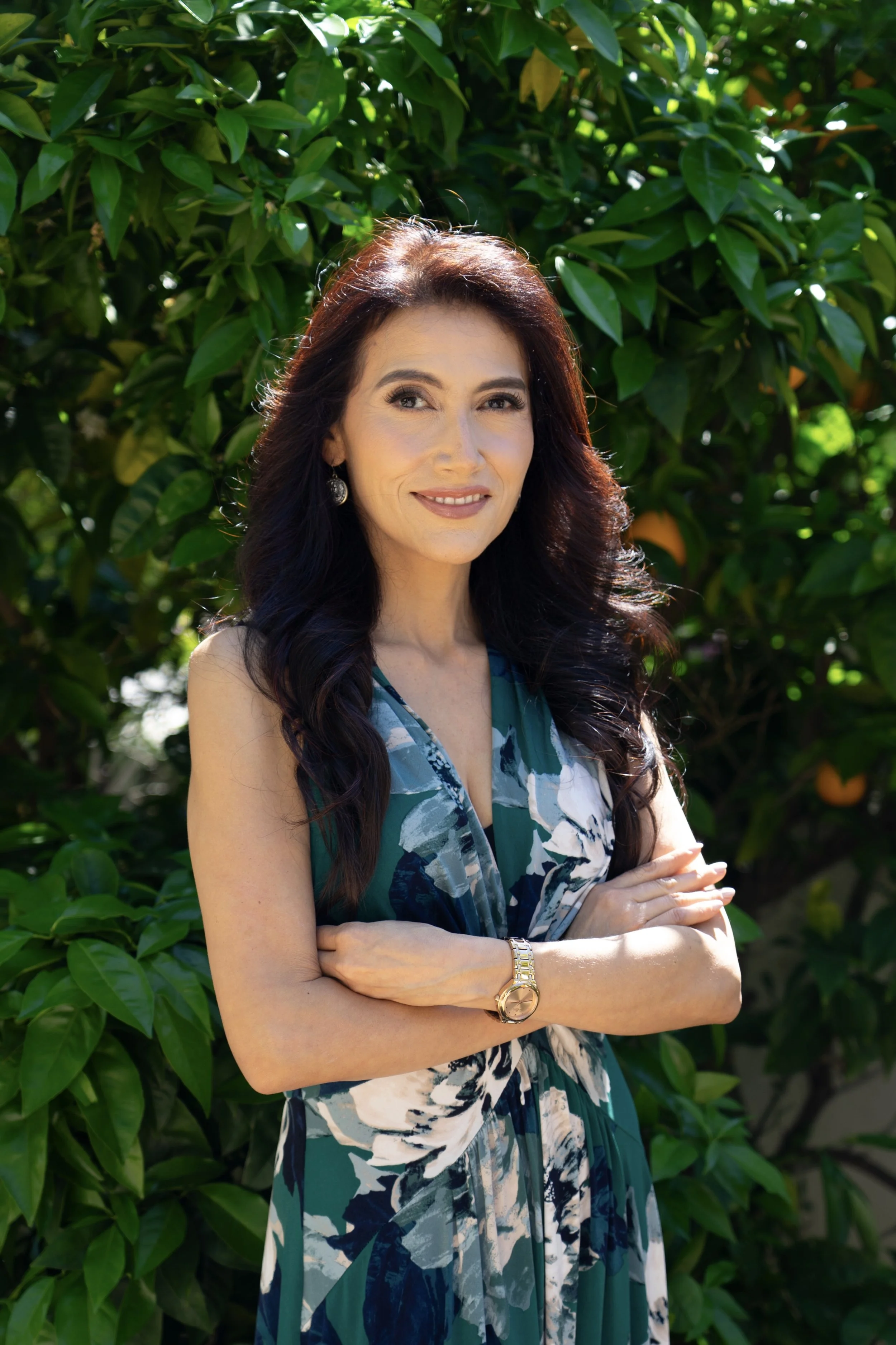 Dr. Janice Sullivan with long dark hair, wearing earrings and a wristwatch, stands outdoors with her arms crossed, in front of lush green foliage, smiling slightly and looking to her left.