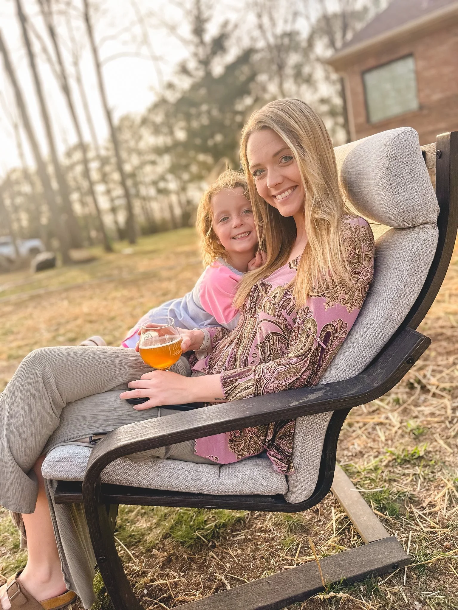 A young woman and a little girl sitting outdoors on a reclining chair, smiling at the camera. The woman is holding a glass of beer, and the girl is leaning on her shoulder in a sunny yard with trees and a house in the background.