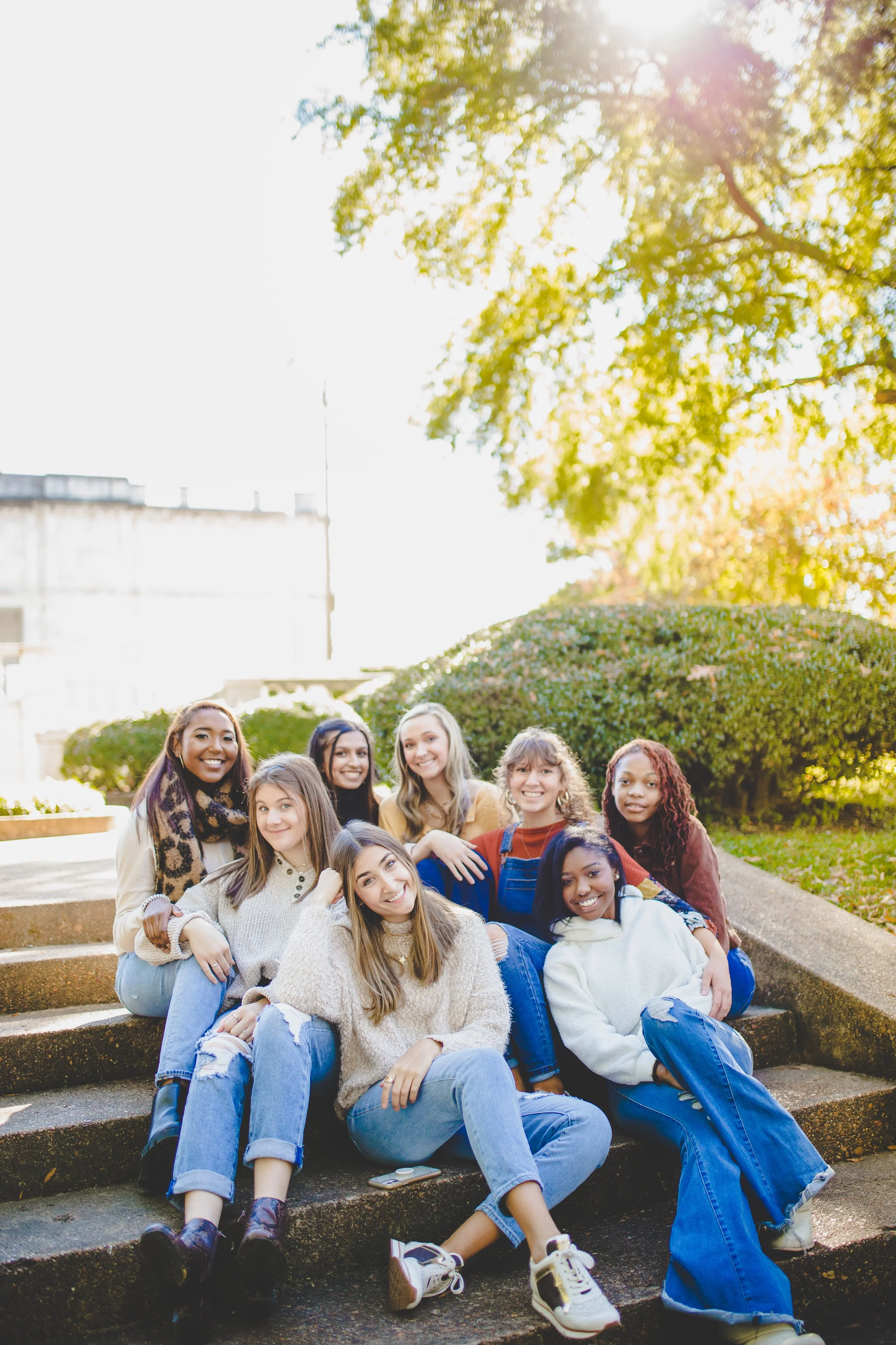 Group of nine young women sitting on outdoor steps in a park during daytime, smiling with sunlight filtering through trees.