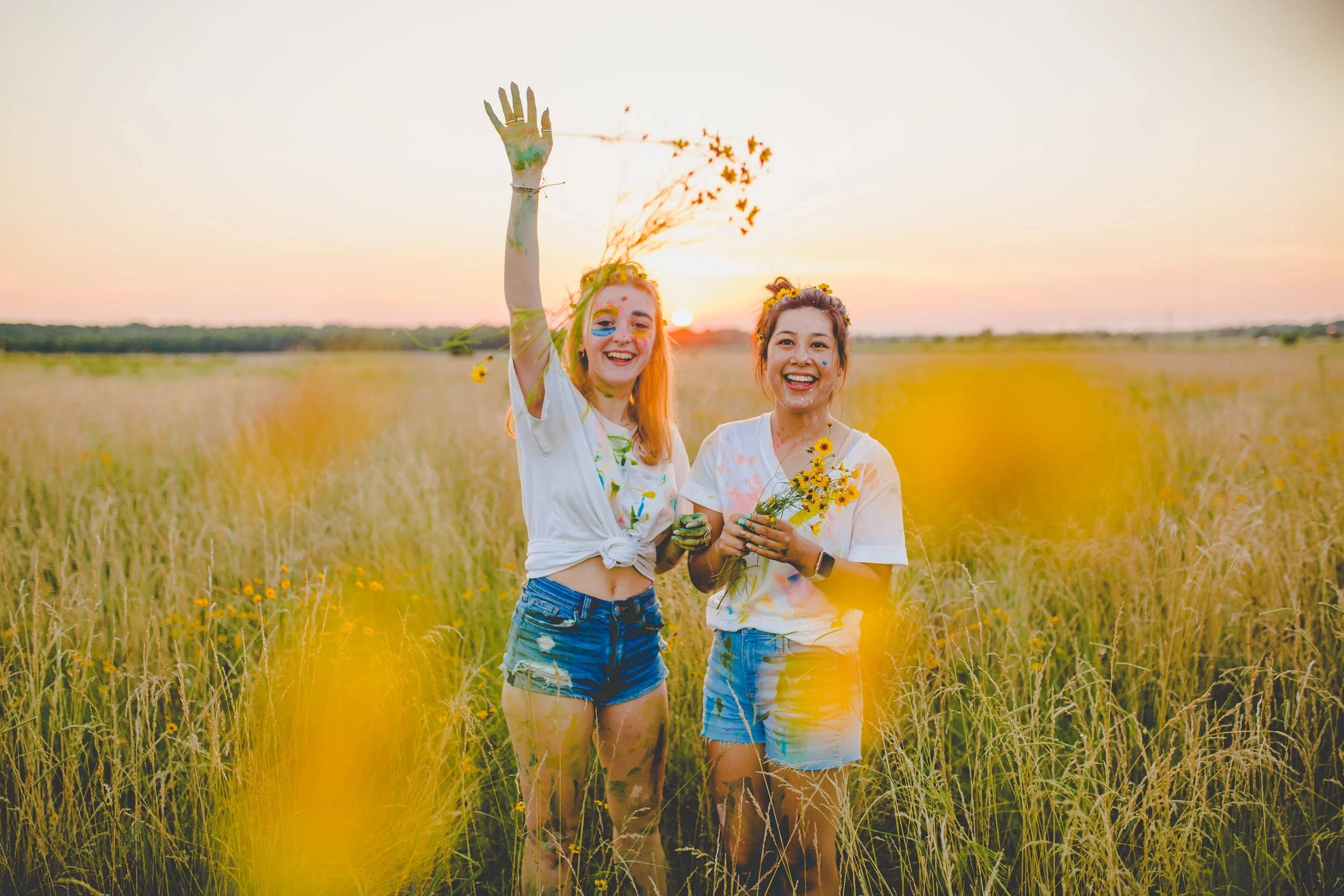 Two women smiling in a field of tall grass at sunset, holding flowers, with paint on their faces and clothes, celebrating outdoors.