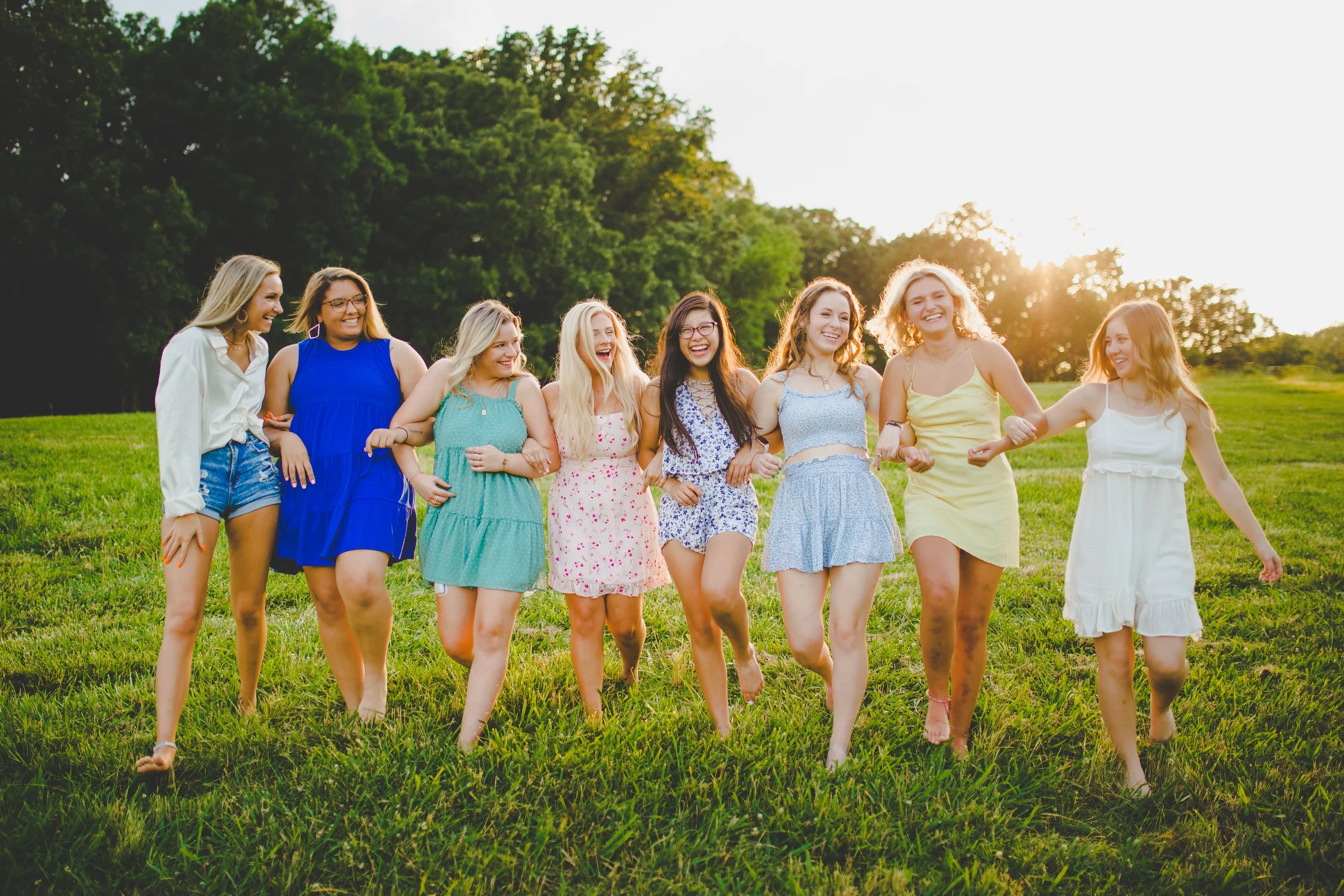 Group of nine young women holding hands, walking on grass in a park at sunset, smiling and laughing.