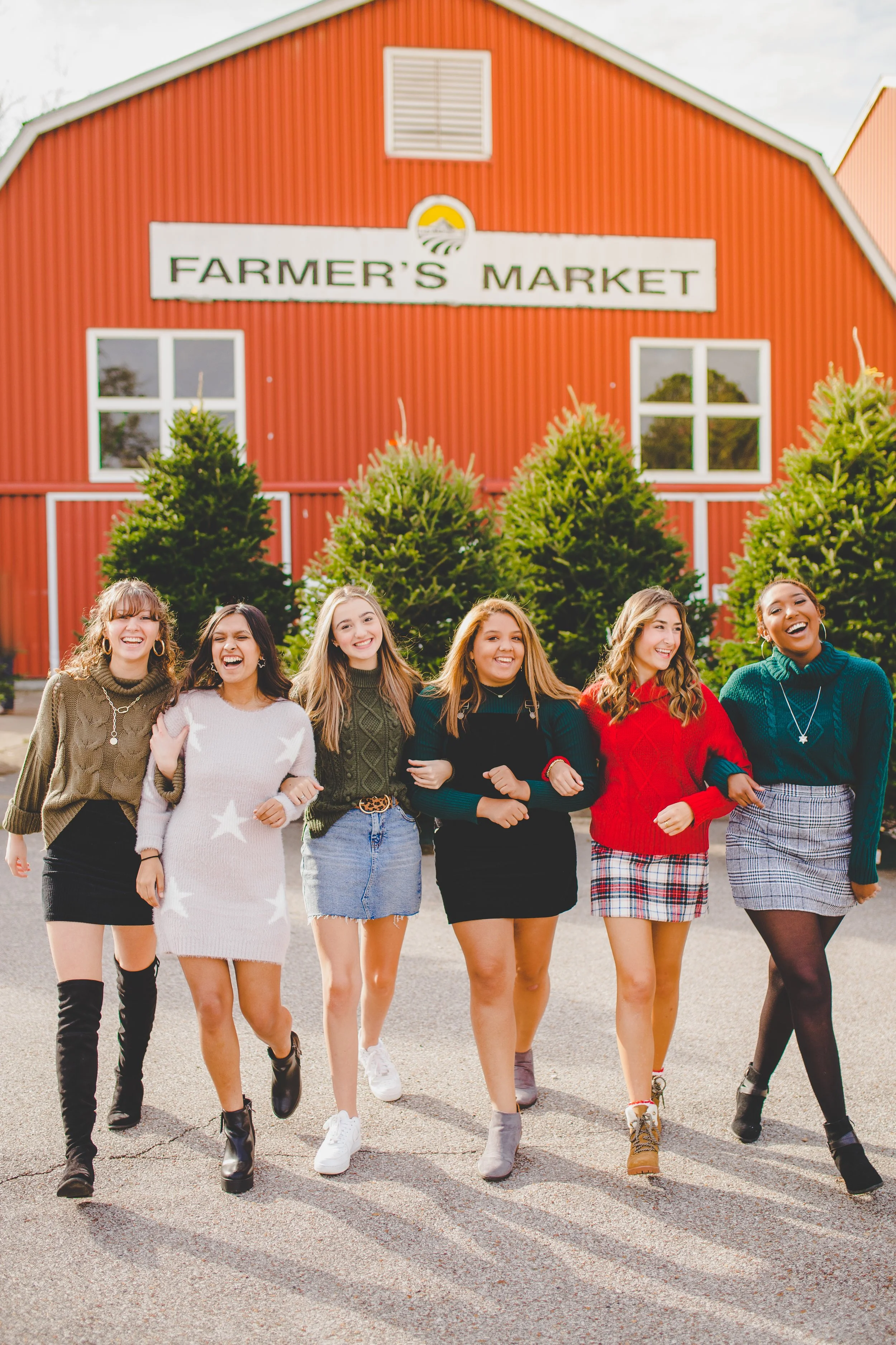 Group of six young women walking arm-in-arm in front of a red barn with a Farmer's Market sign, dressed in sweaters and skirts, smiling and enjoying each other's company.