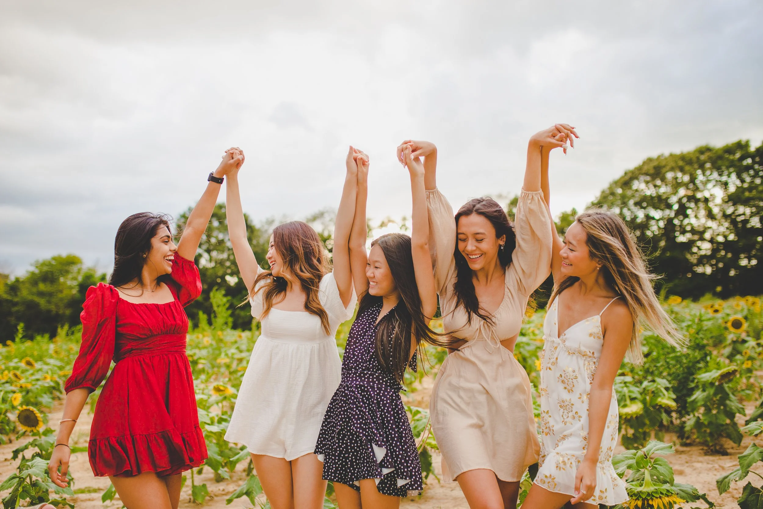 Five women in dresses smiling and holding their hands up in the air, walking through a sunflower field on a cloudy day.
