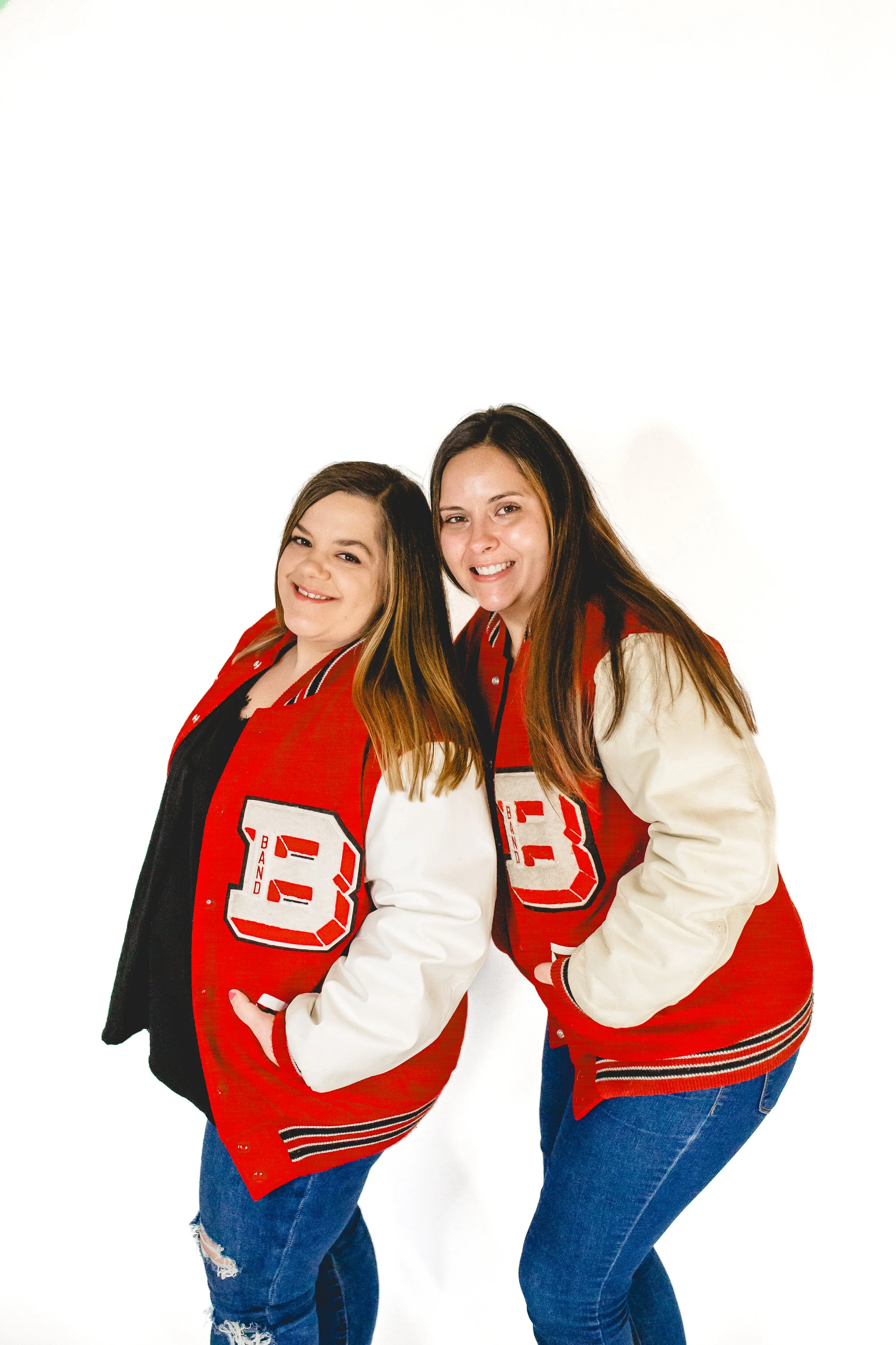 Two women smiling and posing together, wearing red and cream varsity jackets with the letter B on the front, against a white background.