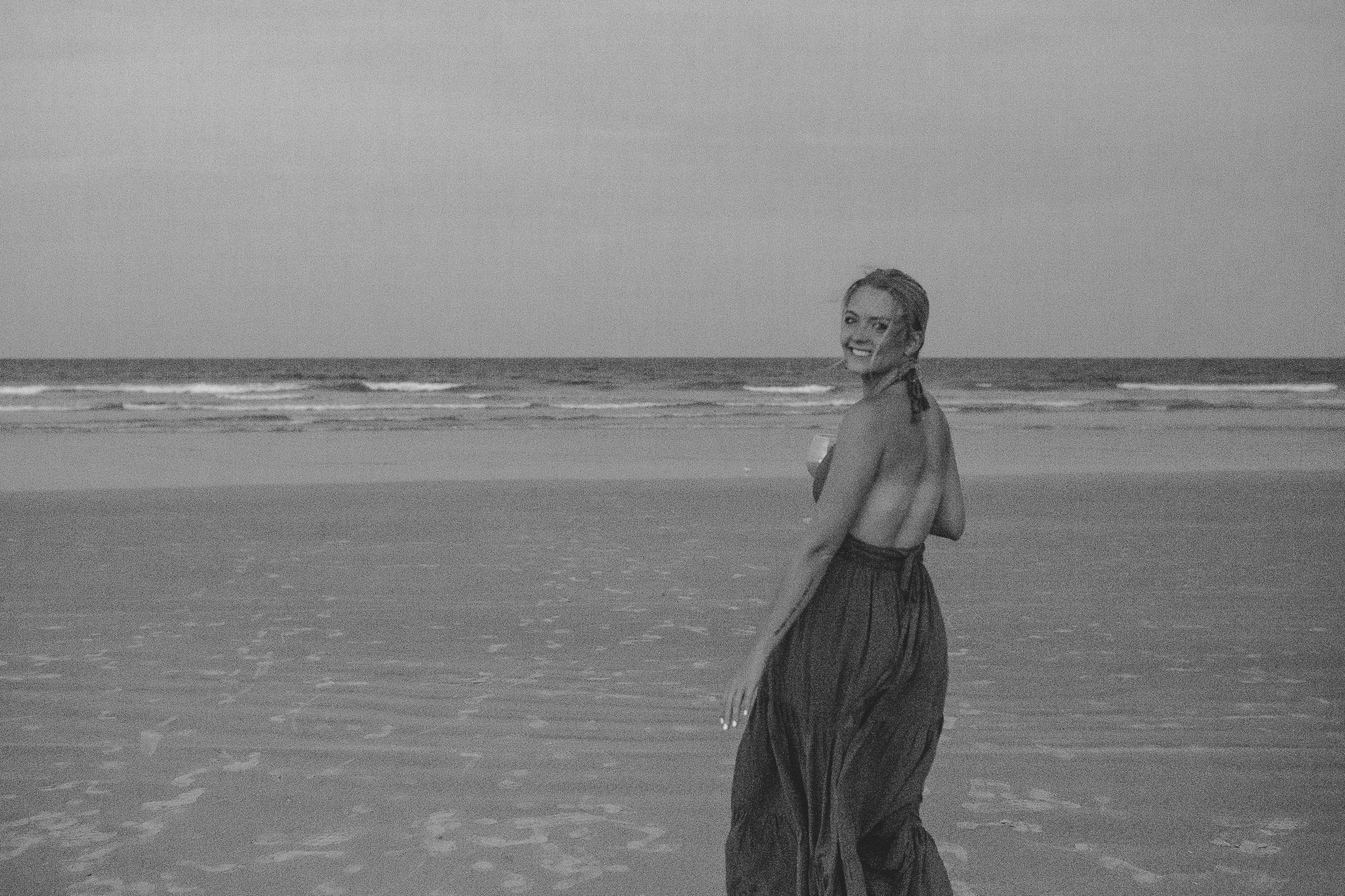 A smiling woman in a strapless dress stands on a beach near the ocean with waves in the background, looking back at the camera.
