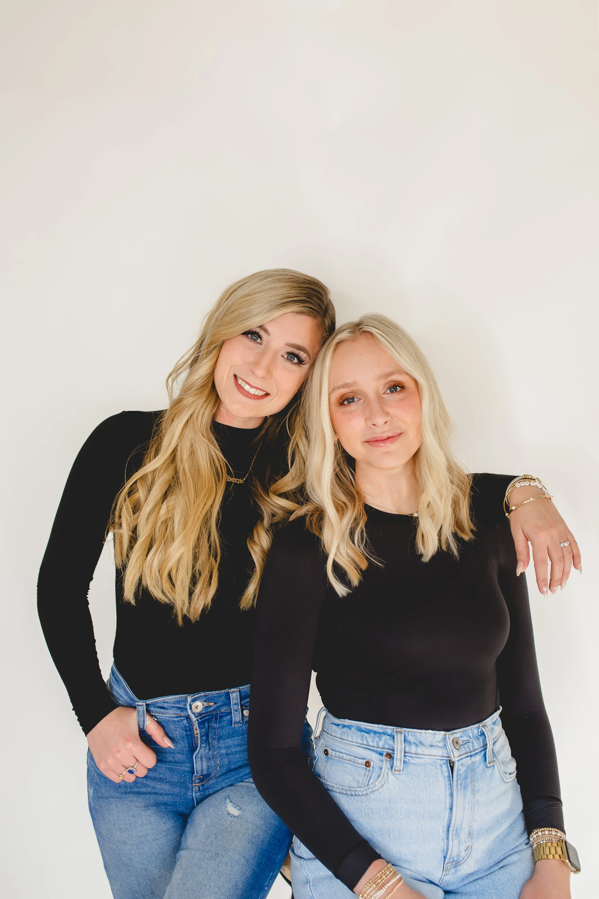 Two women with long blonde hair, wearing black tops and blue jeans, smiling and posing together against a plain white background.