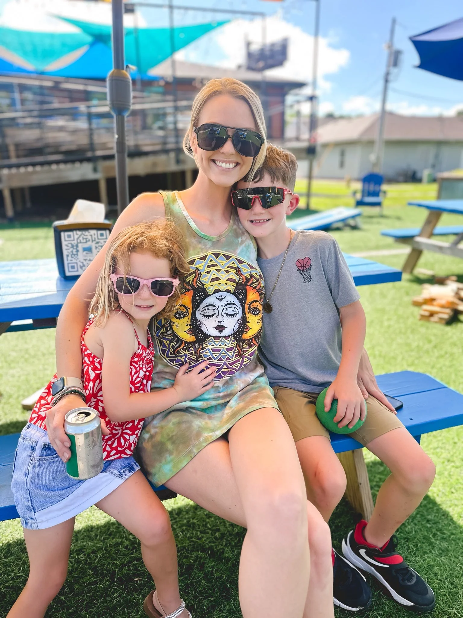 A woman with blonde hair and sunglasses sitting on a blue bench outdoors, smiling with three children, all wearing sunglasses and casual summer clothes, on a sunny day.